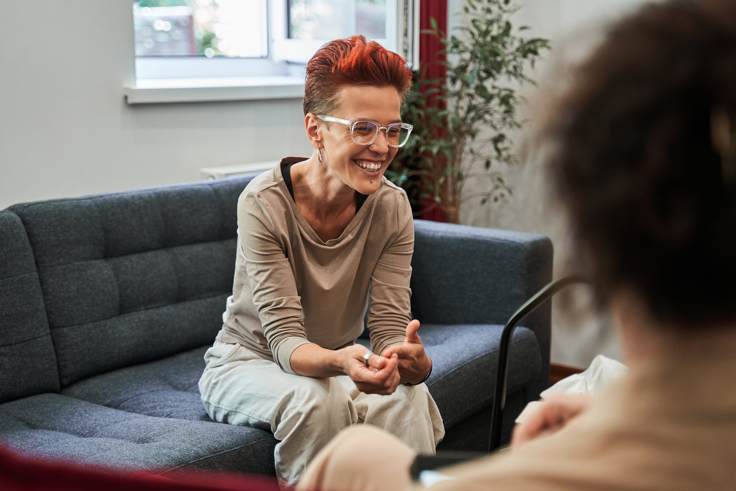 Woman with short red hair and glasses sits on a couch and laughs with a blurry person in the foreground