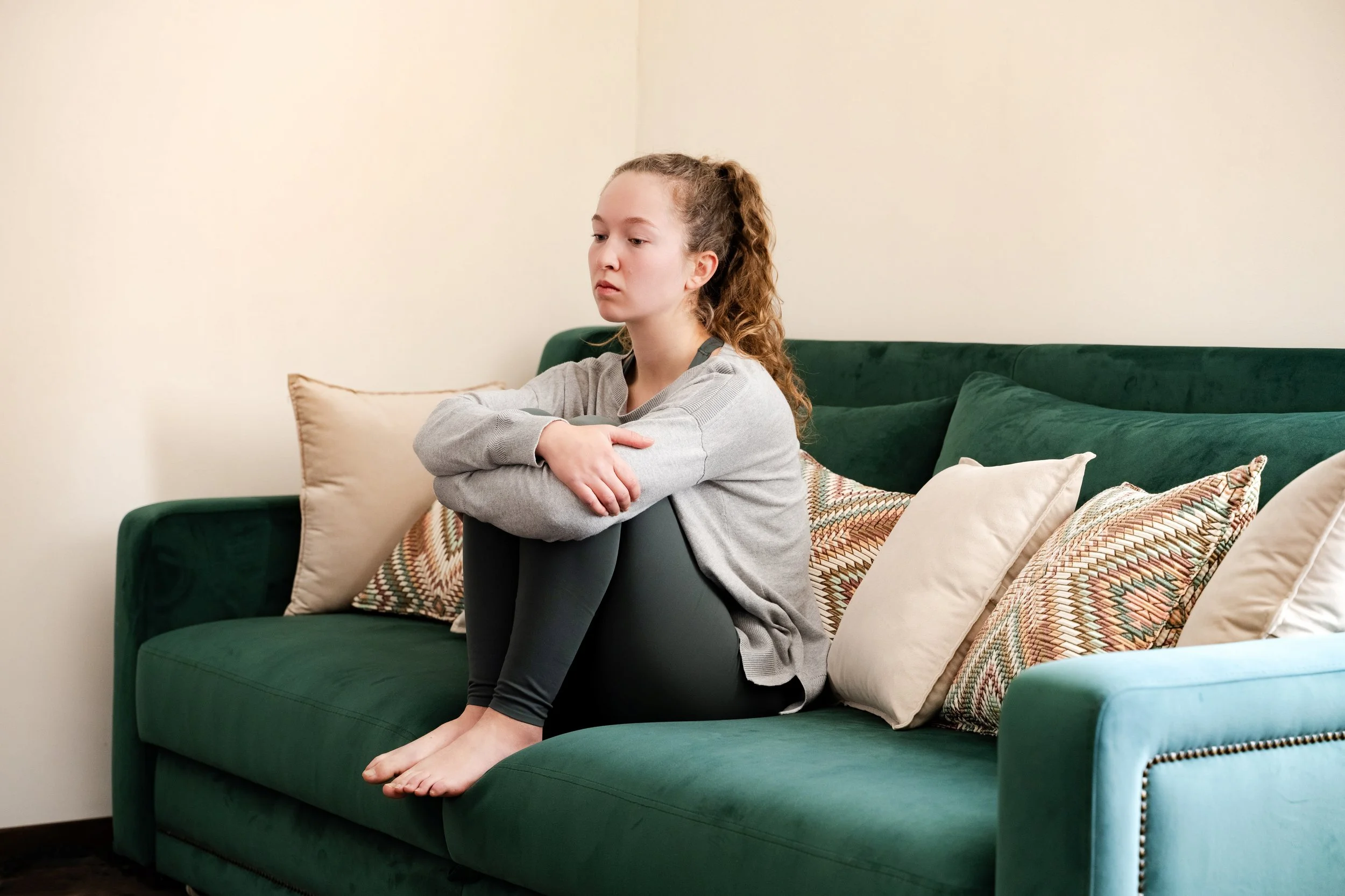 Pensive young woman sitting on a green couch with her legs pulled up and arms resting on her knees