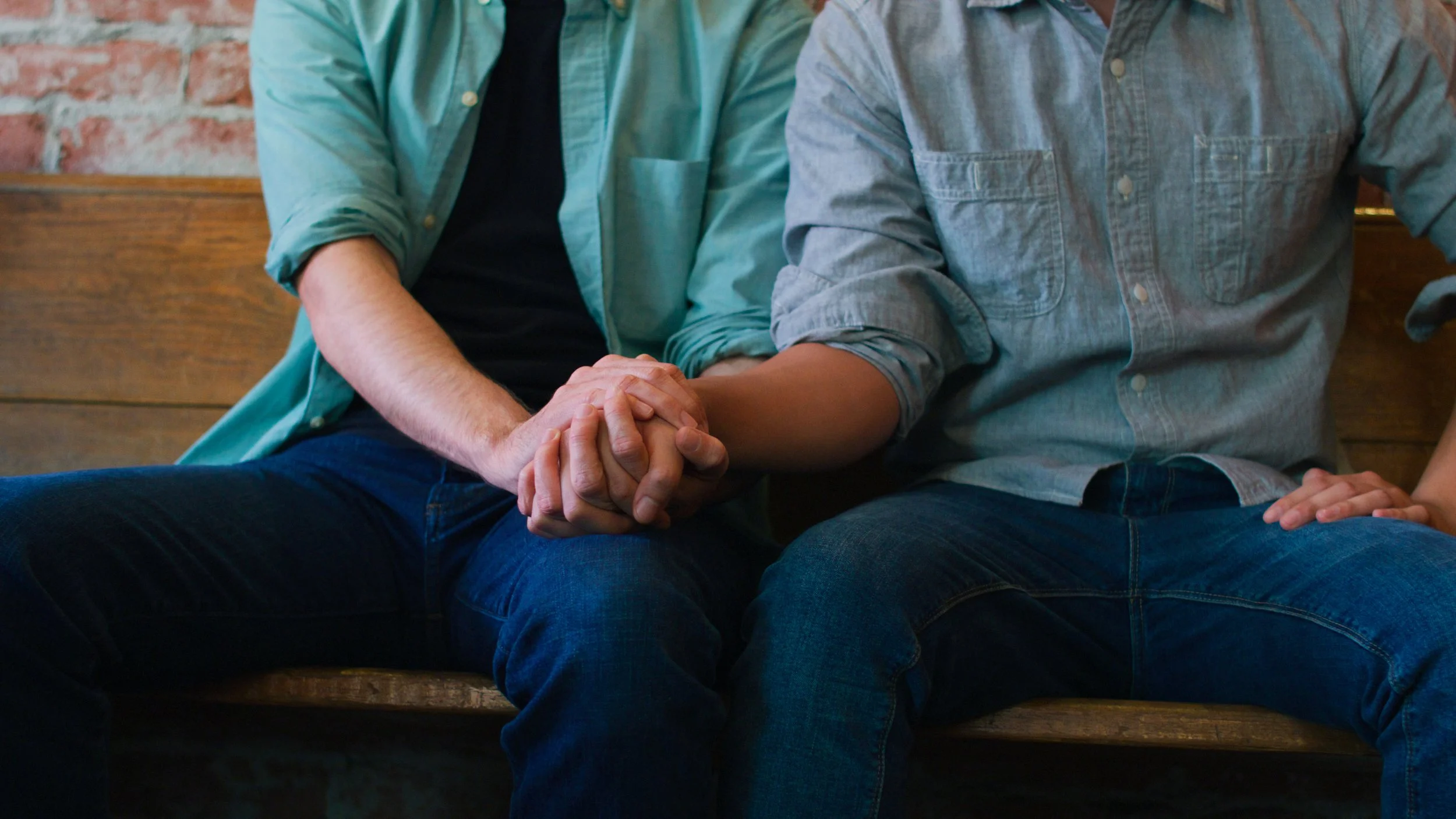 Two men sitting on a bench holding hands, cropped from knees to shoulders