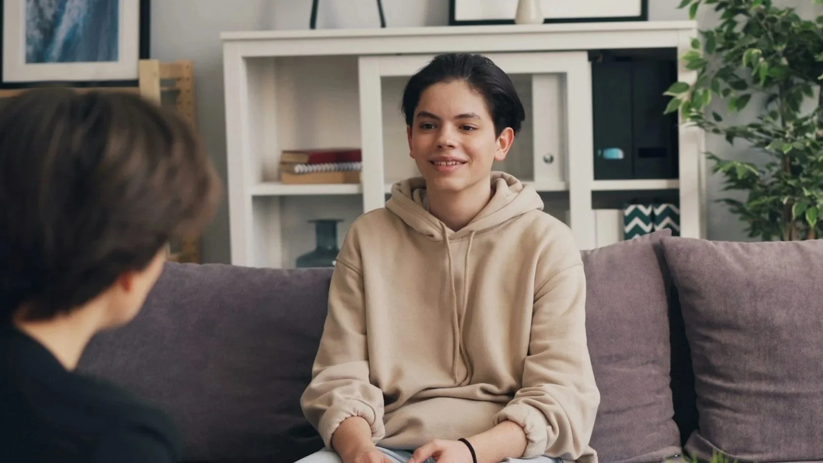 A young man with dark hair and a beige hoodie sitting on a gray couch, talking to another person whose back is turned towards the camera, in a living room with a white bookshelf and plant in the background.