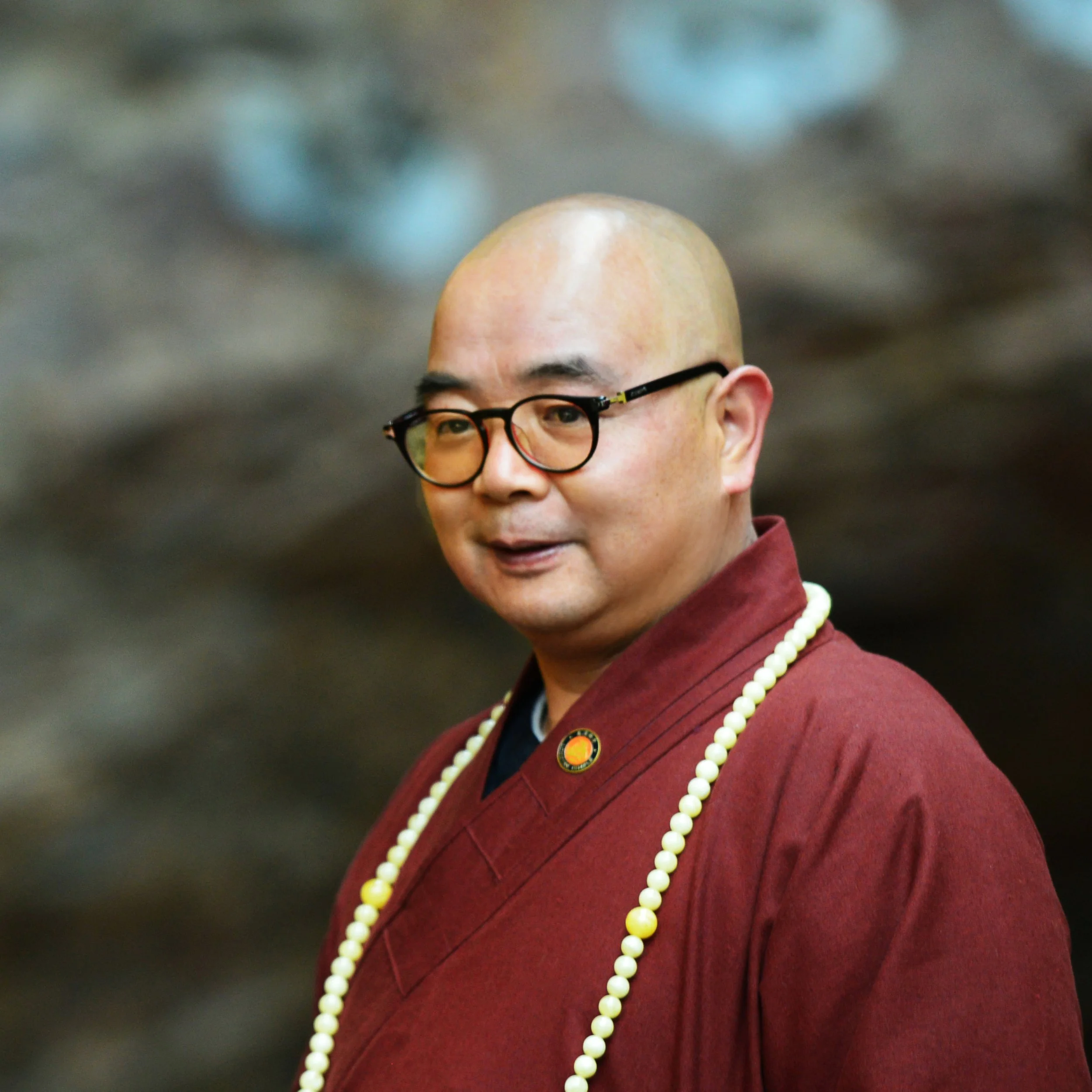 A person with a shaved head wearing glasses, a maroon jacket, and a pearl necklace.