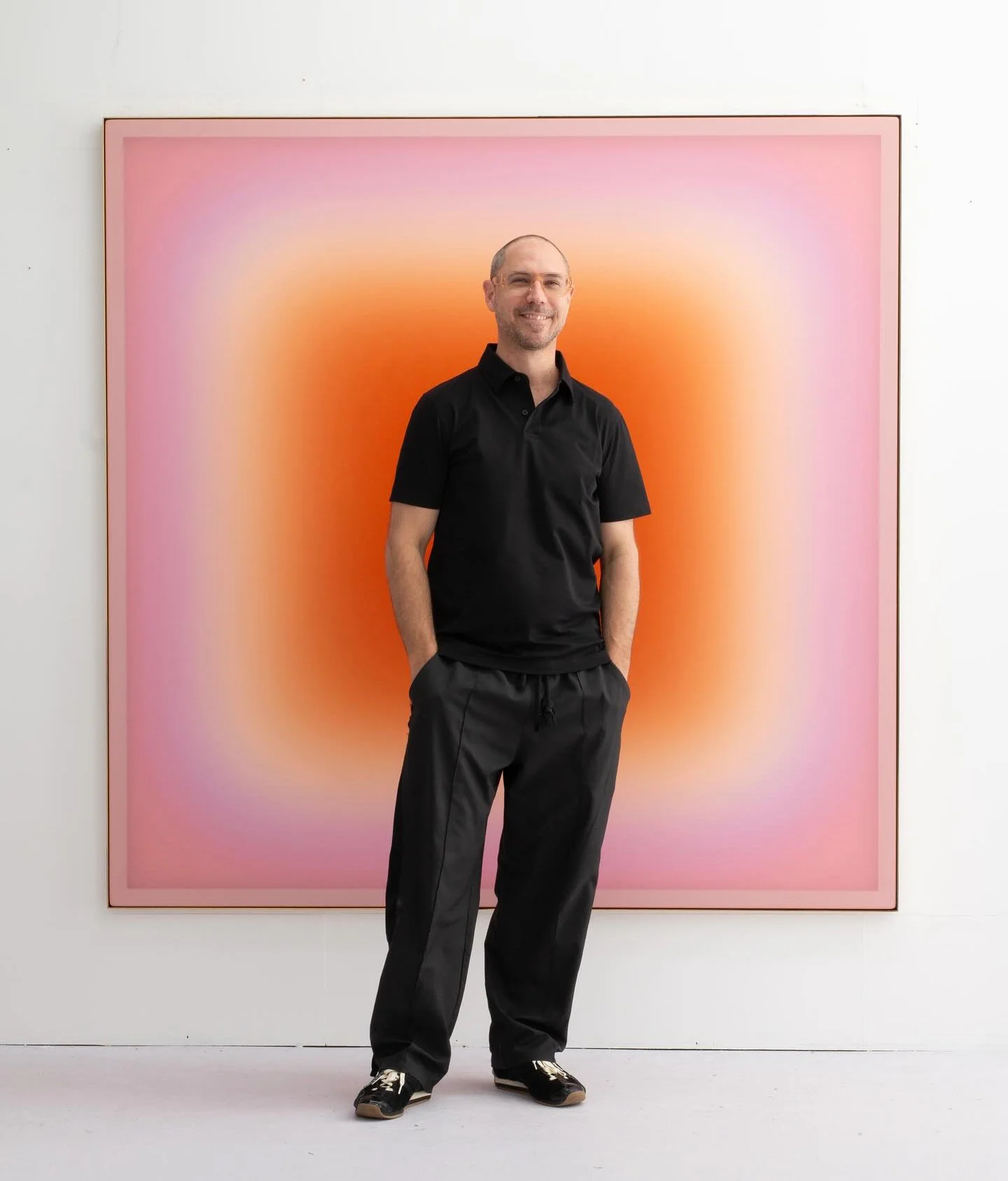 A man standing in front of a colorful, gradient artwork with pink and orange hues, smiling and casually dressed in black clothing.