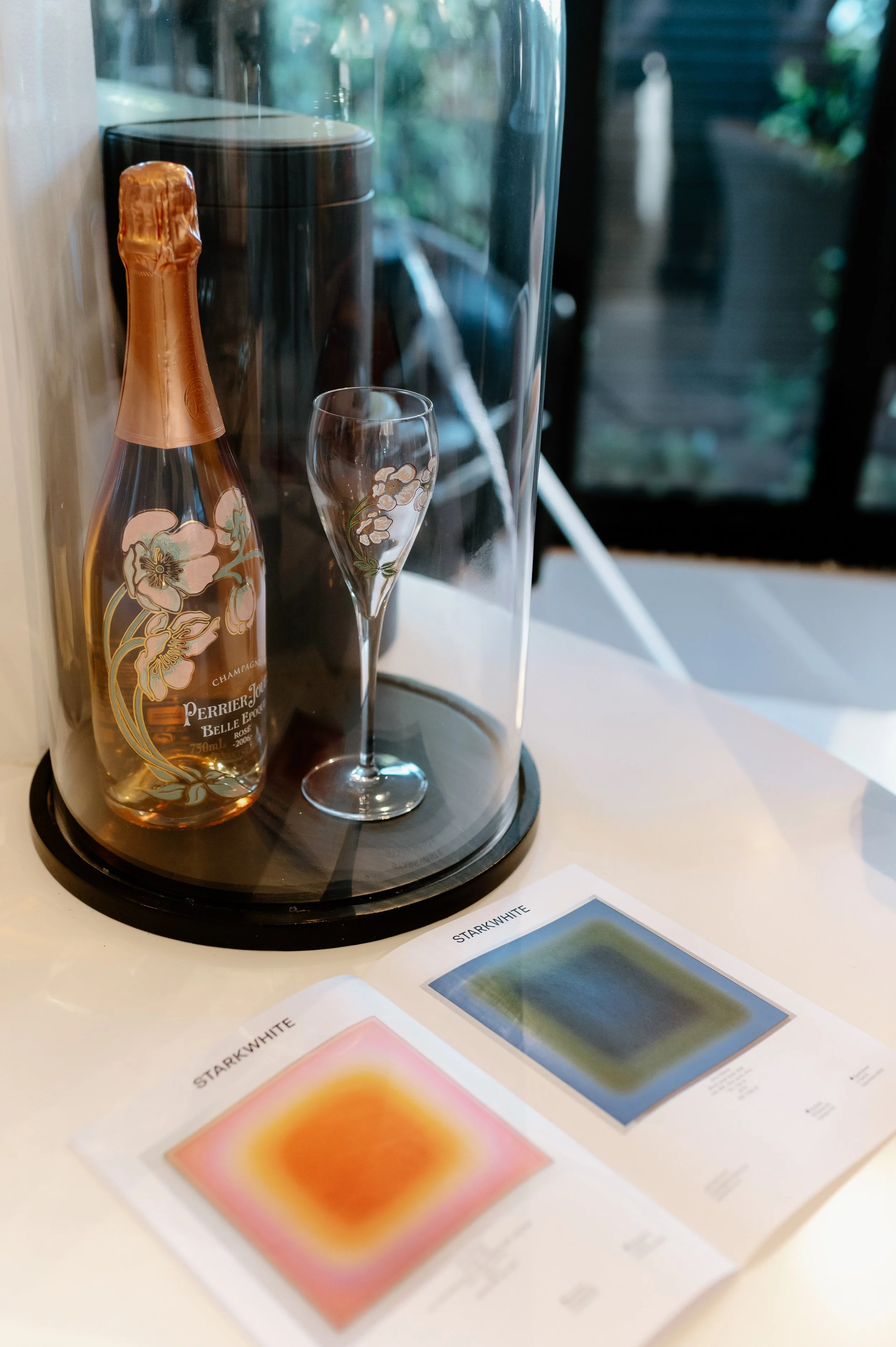 A bottle of Perrier-Jouët Belle Epoque Rosé champagne, a champagne flute with floral decorations, and a black wine cooler on a tray, with colorful color sample cards labeled 'Starkwhite' on the table in front of a window showing outdoor greenery.