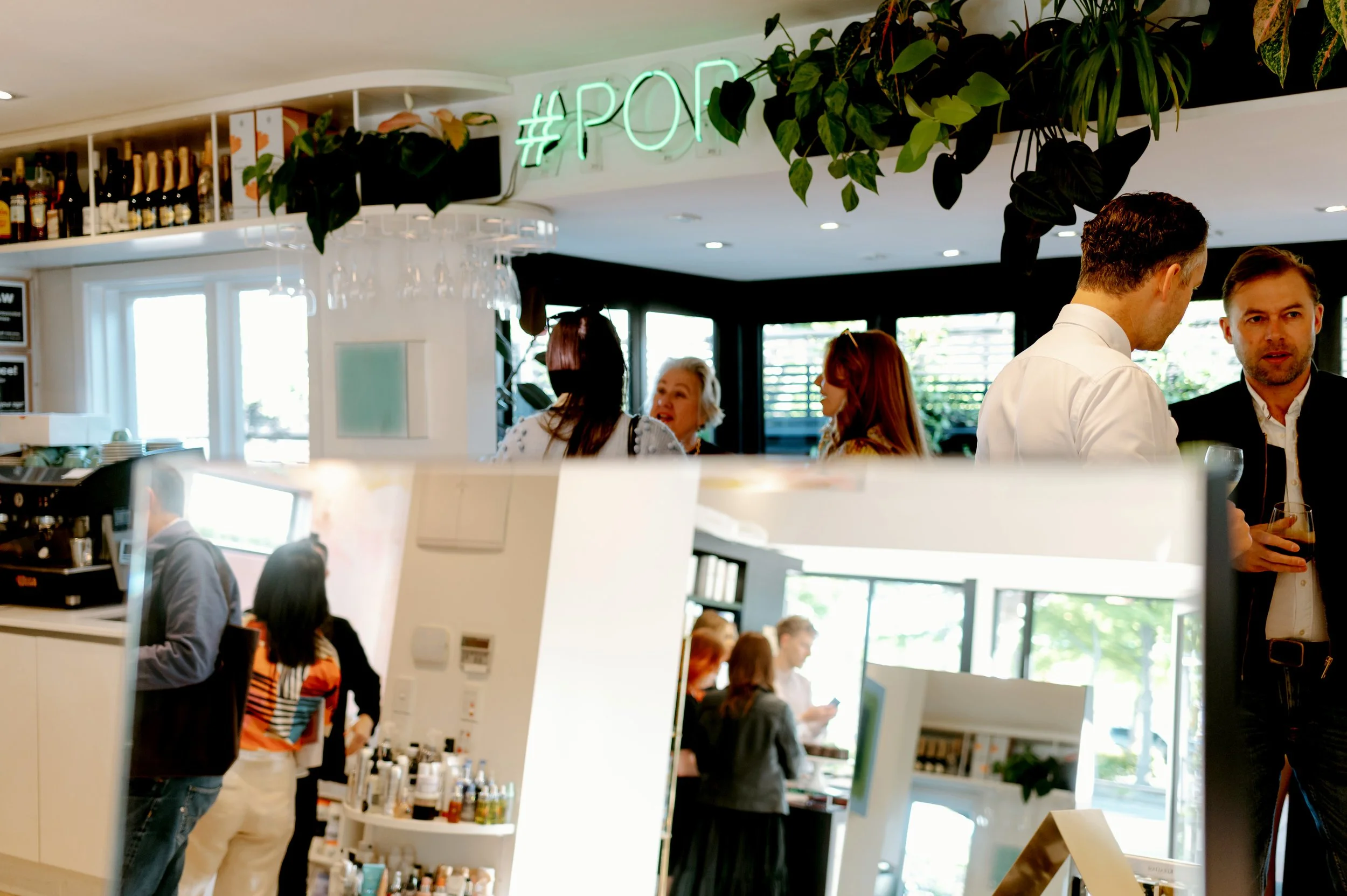 People socializing inside a modern bar or restaurant with a hanging #POP sign and lush greenery on the ceiling.