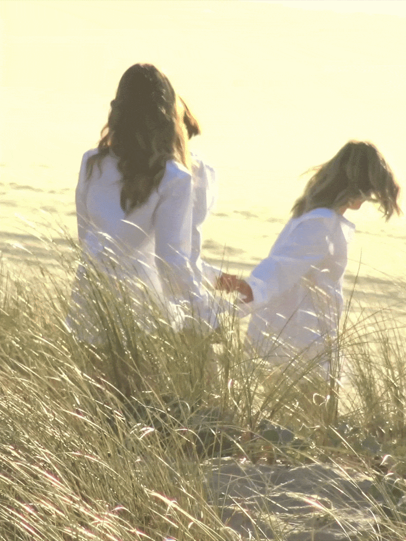 Two women in white shirts walking through tall grass on a beach at sunset, holding hands.