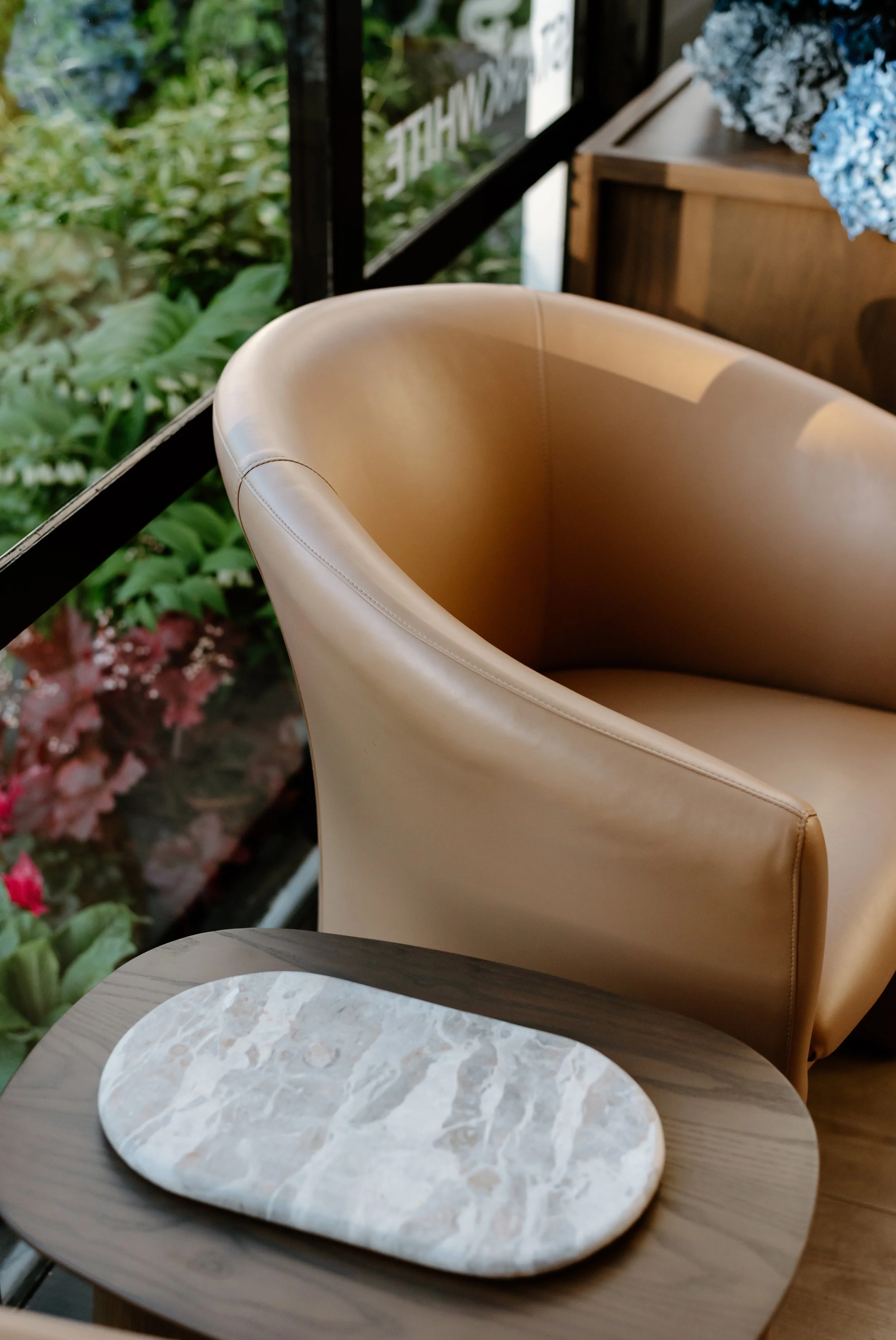 A beige leather chair beside a gray wooden table with a white marble oval tray on top, located near a window with greenery outside.