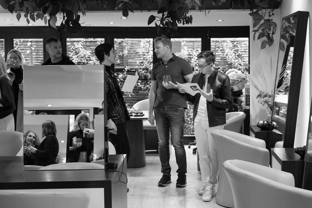 People socializing in a modern indoor space with chairs, a mirror, and large windows with foliage outside.