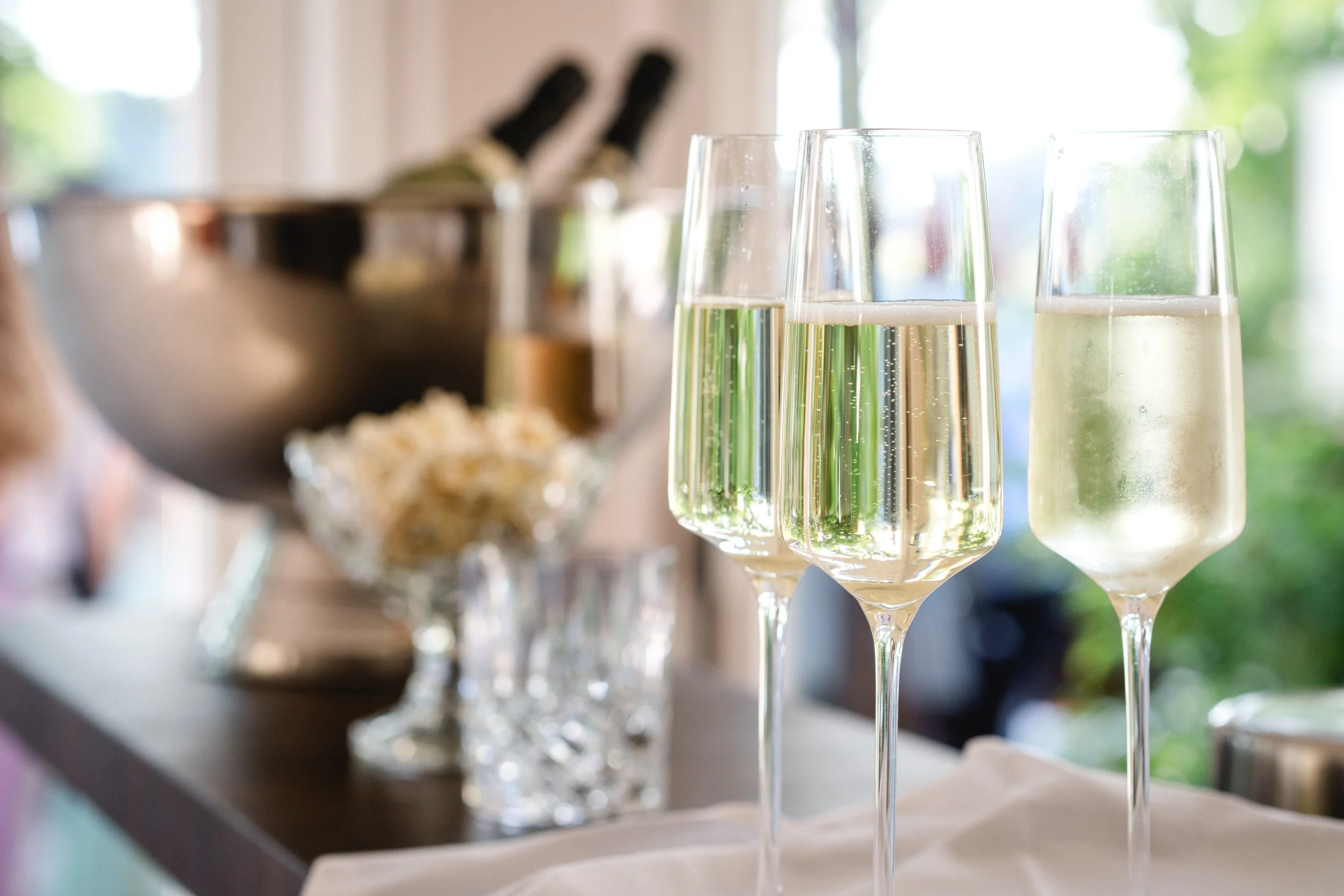 Three champagne flutes filled with sparkling wine on a table. In the background, there is a blurred bowl of snacks and a window with greenery outside.