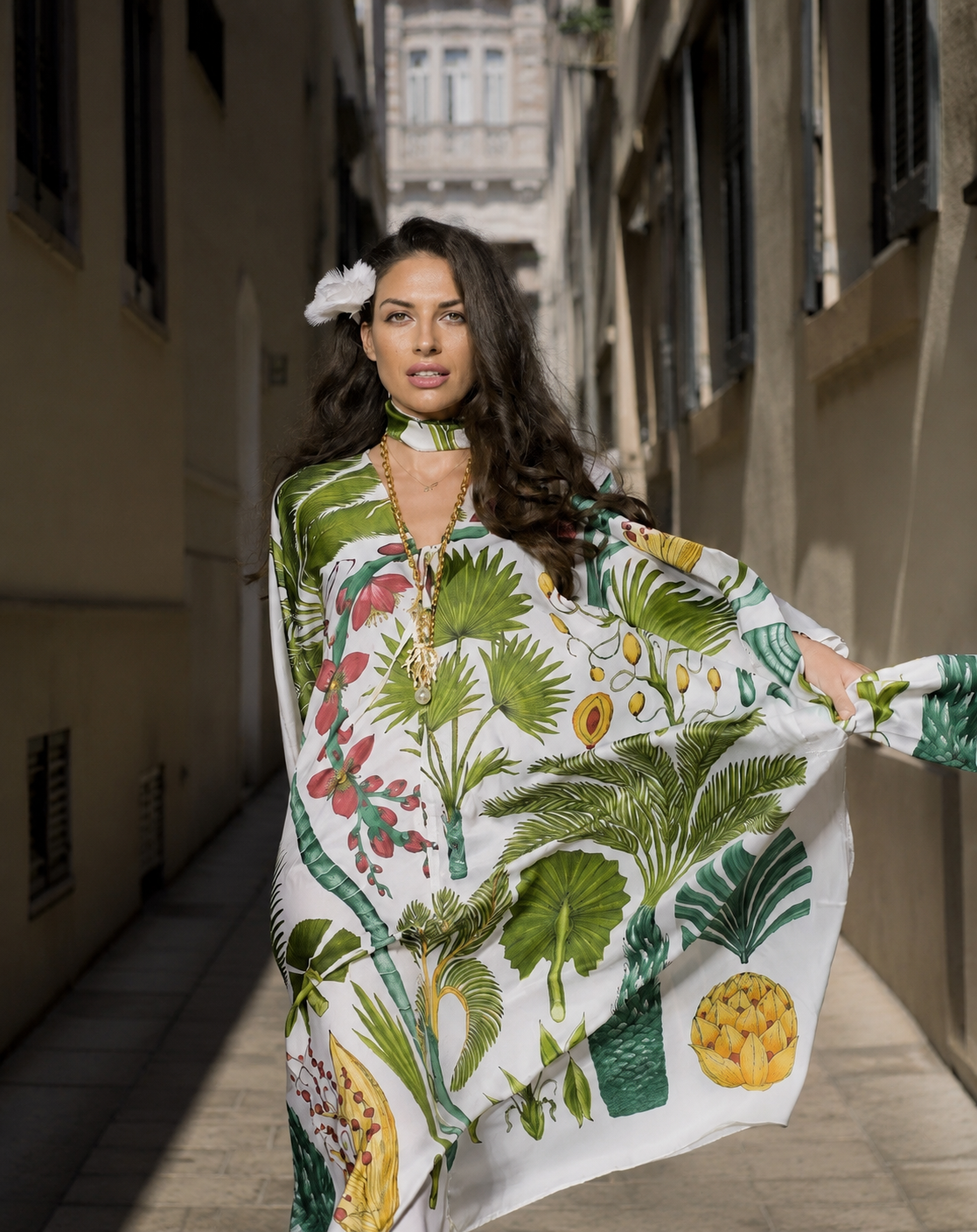 A woman with long dark wavy hair, wearing a large white dress with green tropical leaf and fruit prints, standing in an alleyway with tall buildings on either side.