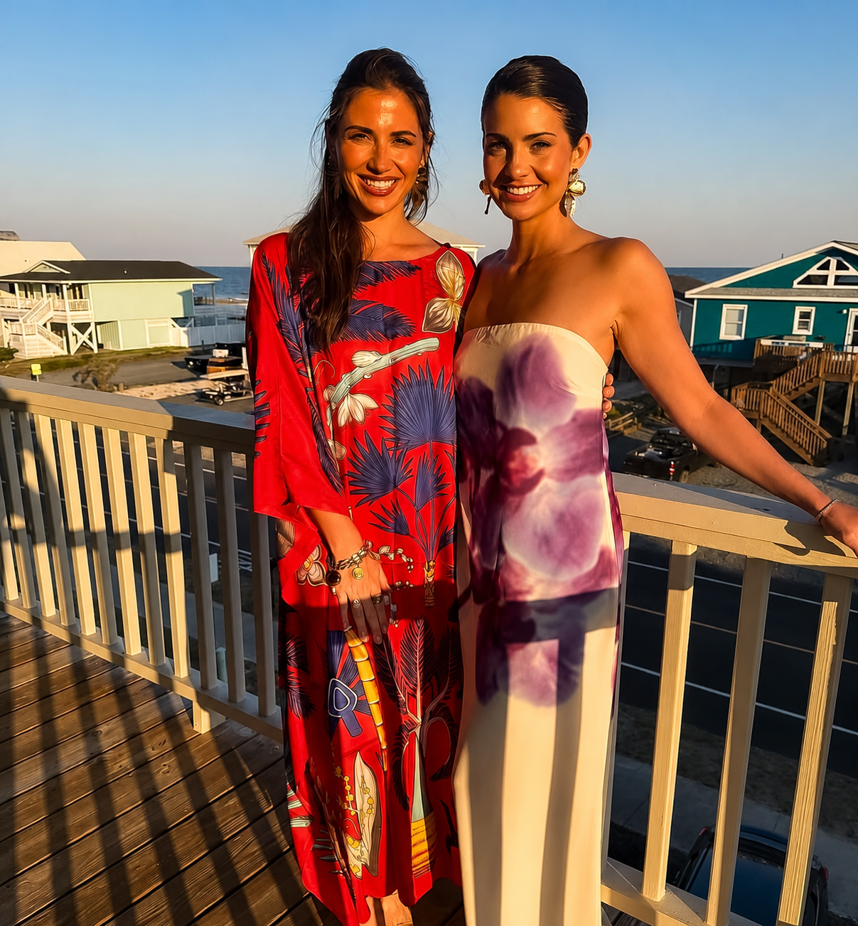 Two women standing on a balcony near the beach during sunset, dressed in colorful summer dresses, smiling at the camera.