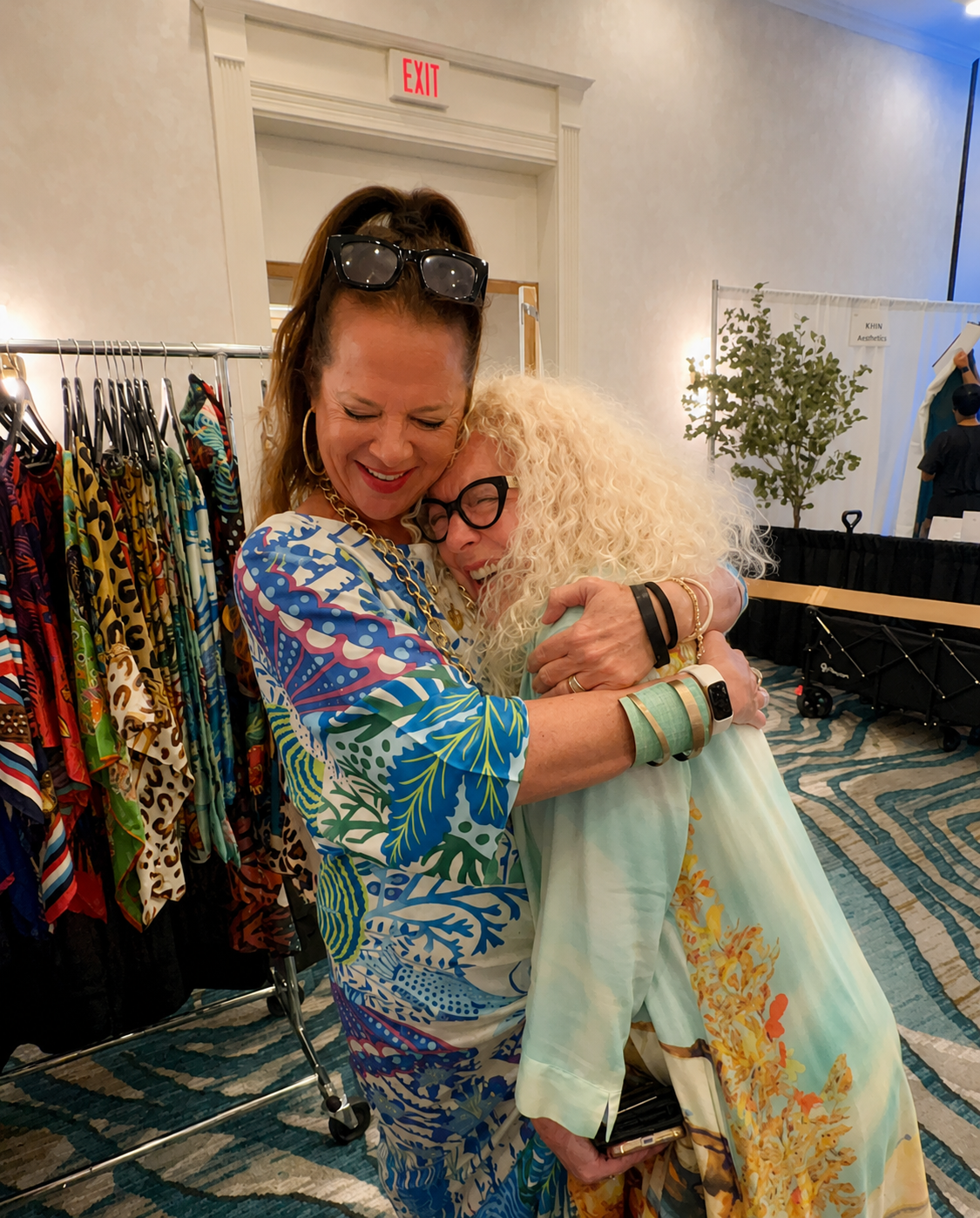 Two women hugging and smiling at a colorful clothing store or boutique, with racks of vibrant garments in the background.