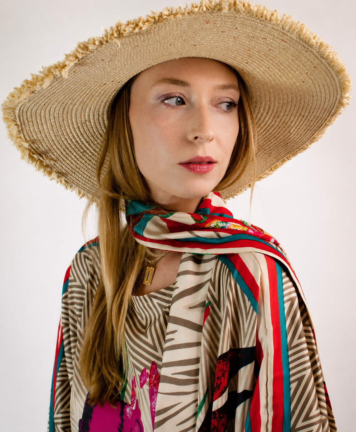 Woman with long hair wearing a large straw hat with frayed edges and a colorful, patterned scarf around her neck, dressed in a bold, patterned top.
