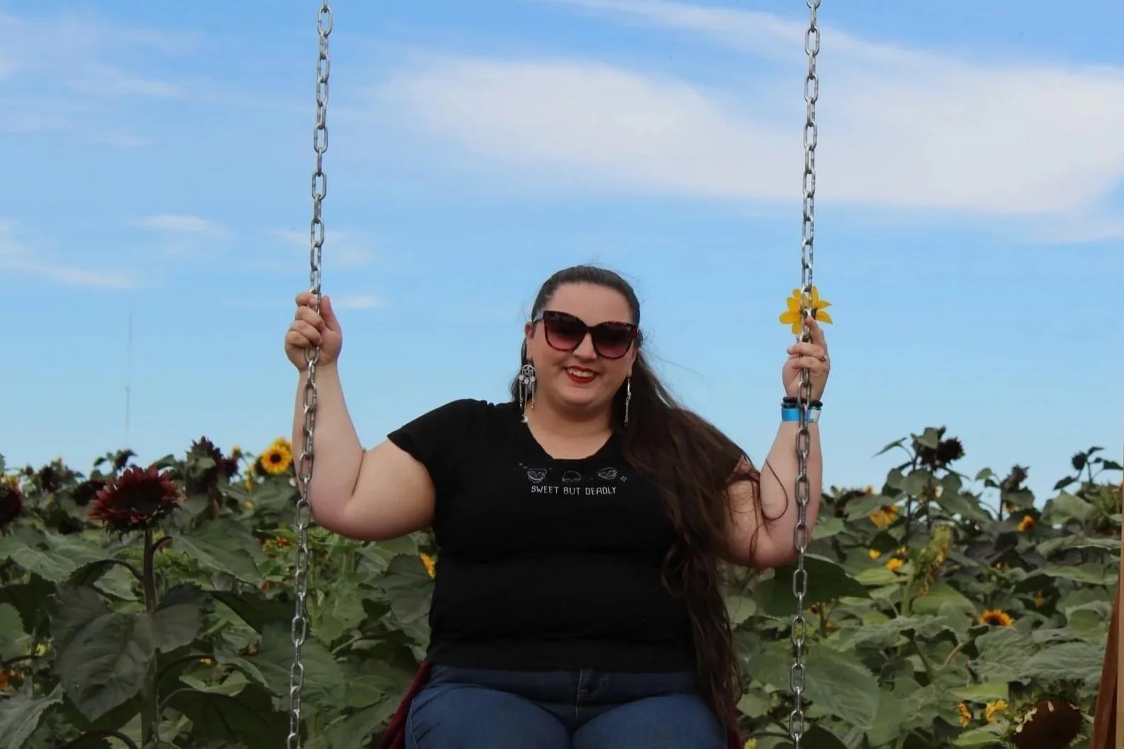 A smiling woman wearing sunglasses and earrings sitting on a swing amidst a sunflower field with a blue sky background.