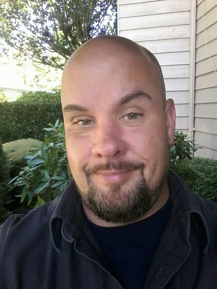 A man with a goatee and shaved head smiling in an outdoor setting with greenery and a house siding in the background.