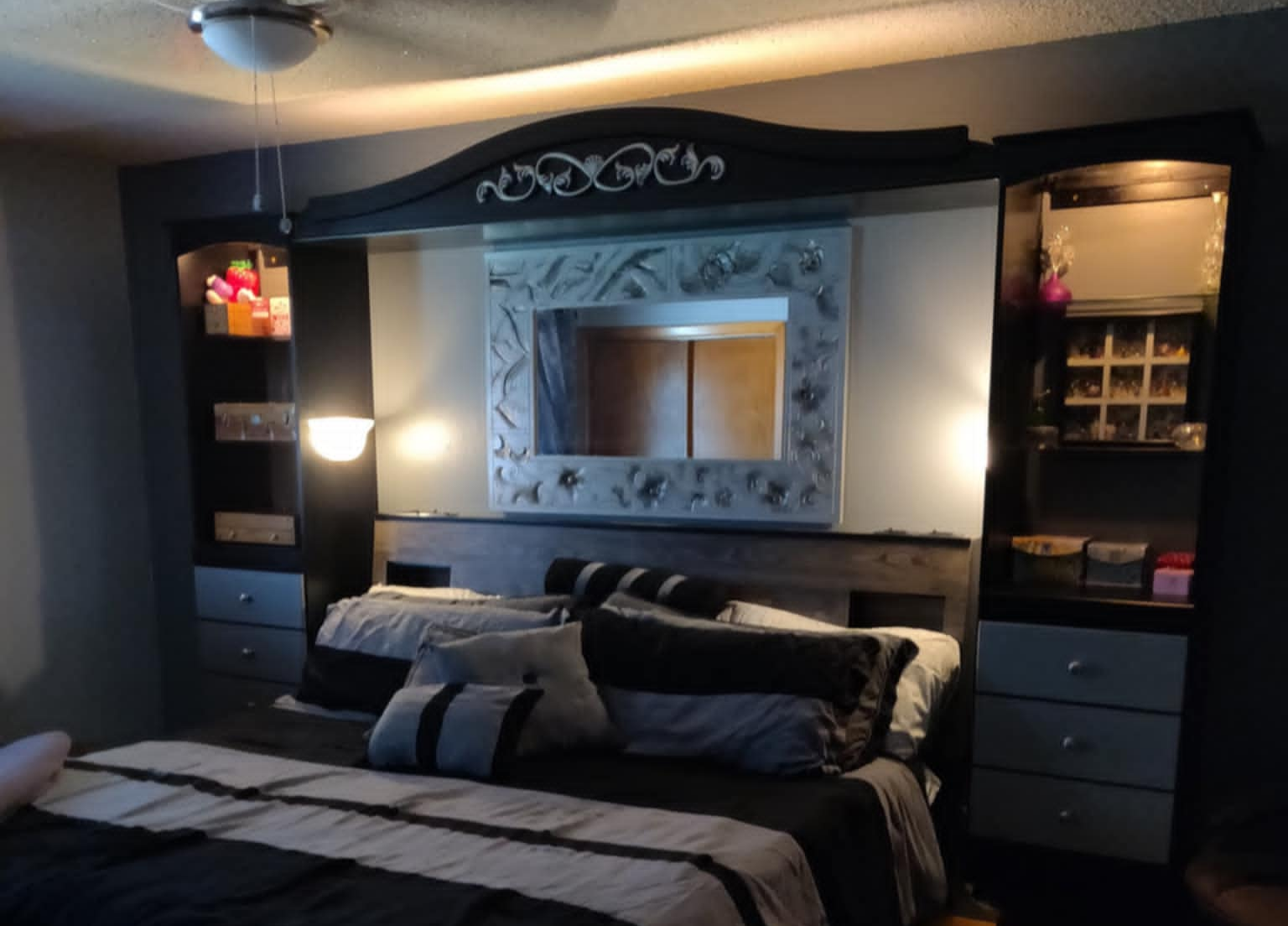 A bedroom with a bed, a large mirror with decorative frame, black furniture, and shelves with various items.