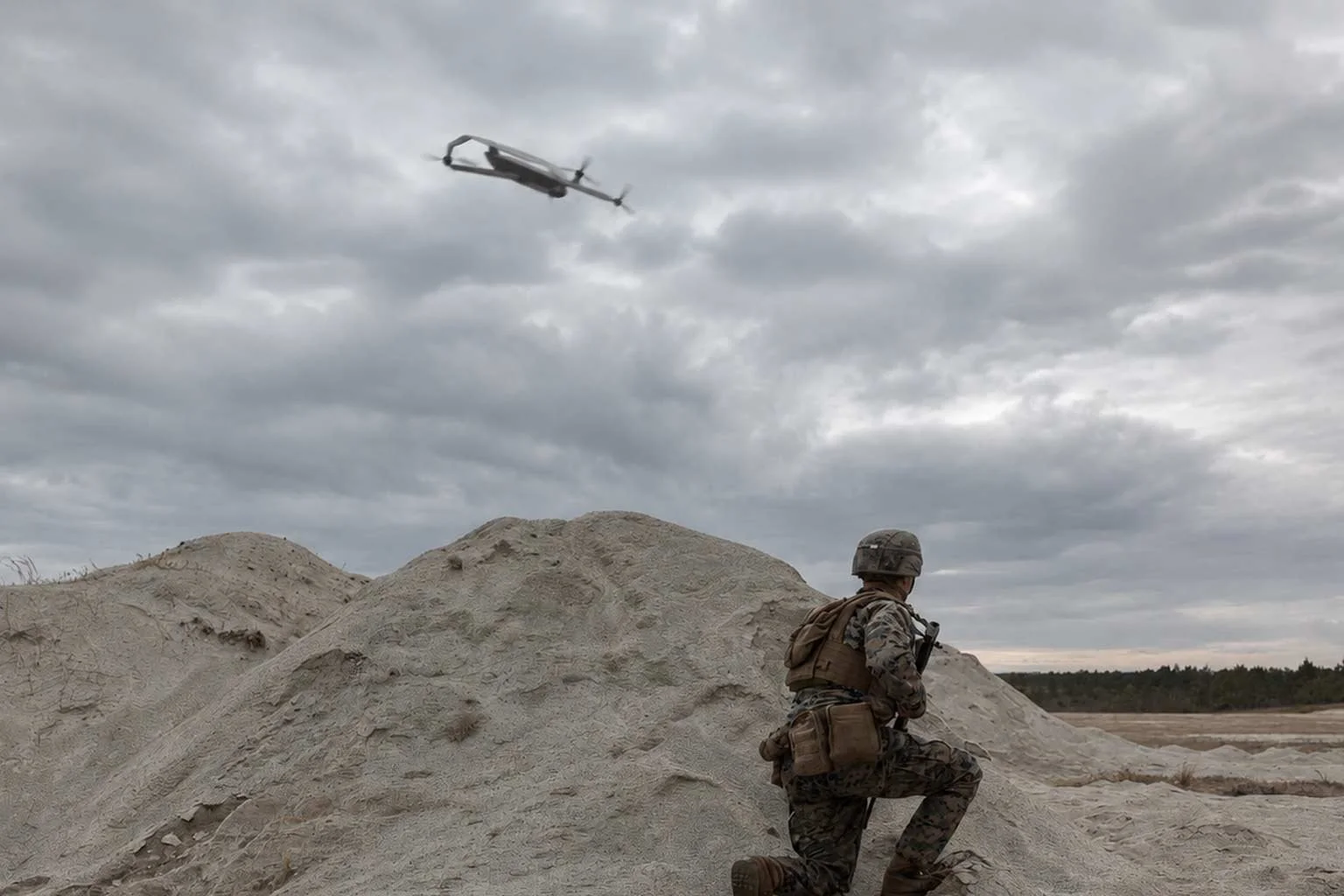 A soldier in camouflage uniform kneeling on a sandy terrain, holding a weapon, with a military helicopter flying in a cloudy sky above.