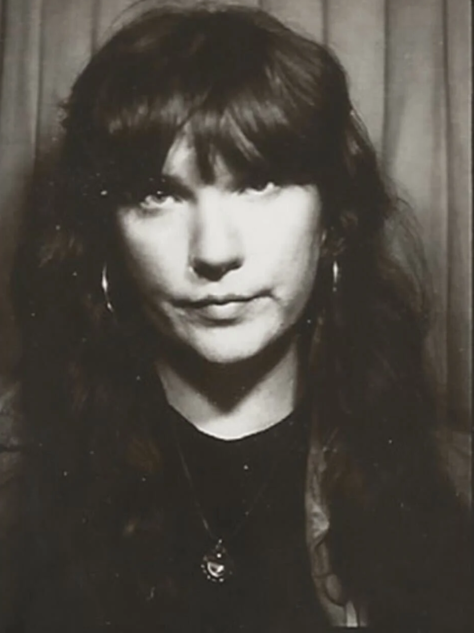 Black and white photo of a woman with long hair, hoop earrings, a necklace, and a septum piercing, standing in front of a wooden background.