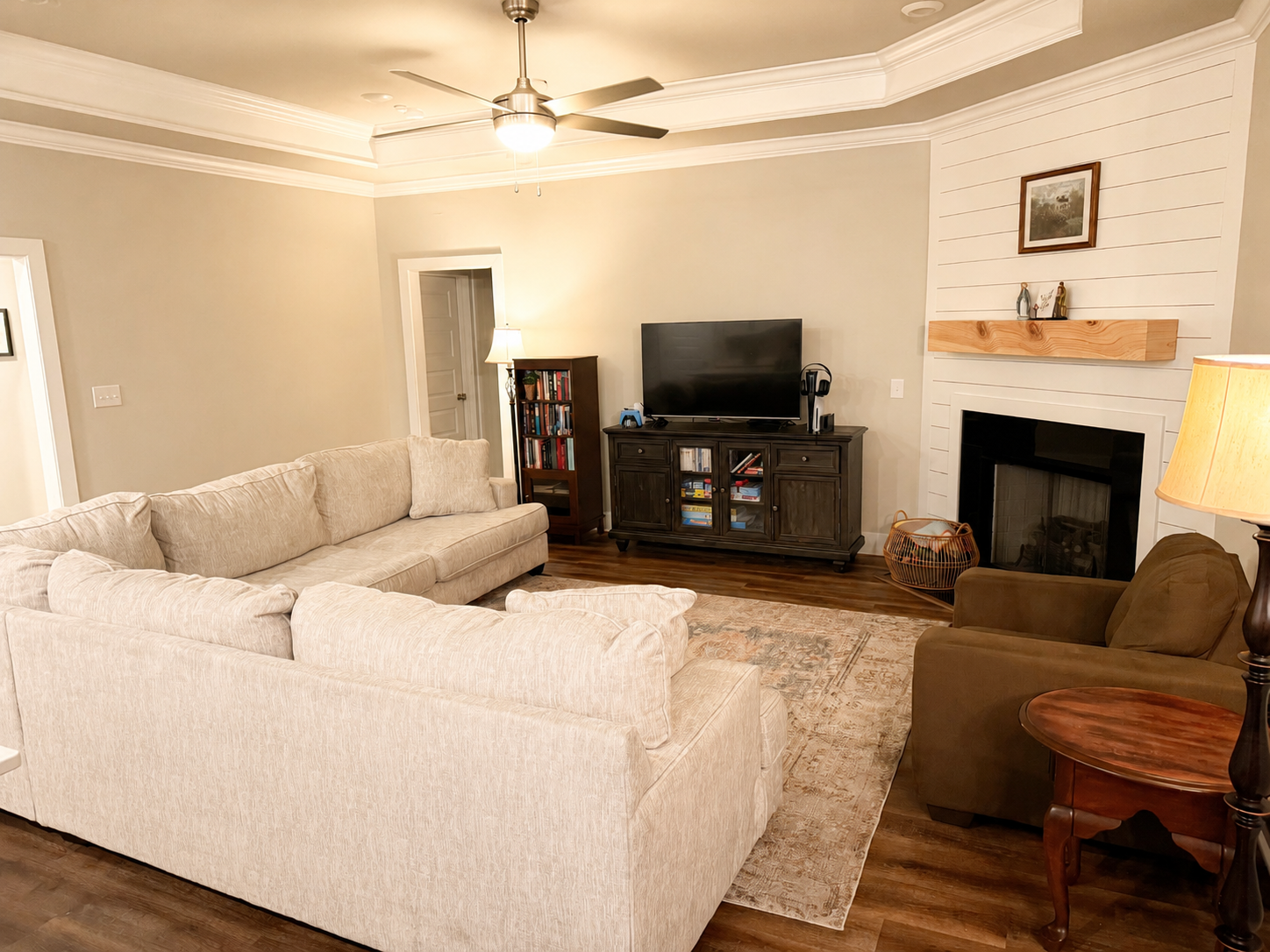 Living room with beige sofa set, brown armchair, wooden side table, white shiplap wall with fireplace, black TV on dark wood cabinet, bookshelf, ceiling fan with lights, rug on hardwood floor.