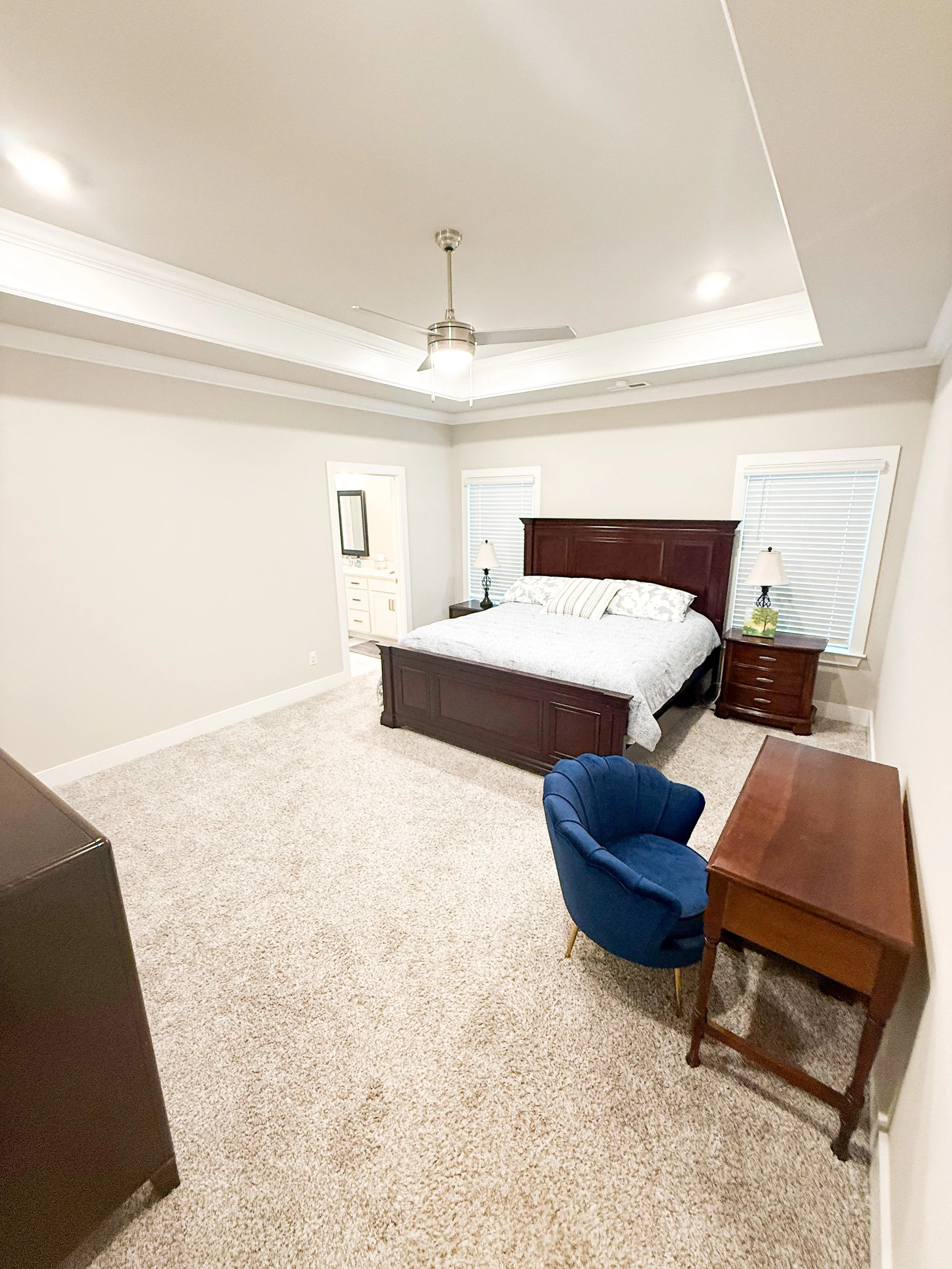 Bedroom with a bed, two nightstands with lamps, a wooden desk, a blue upholstered chair, and a small vanity with a mirror, walls painted in a light neutral color, carpeted floor, ceiling fan with light, and blinds covering two windows.