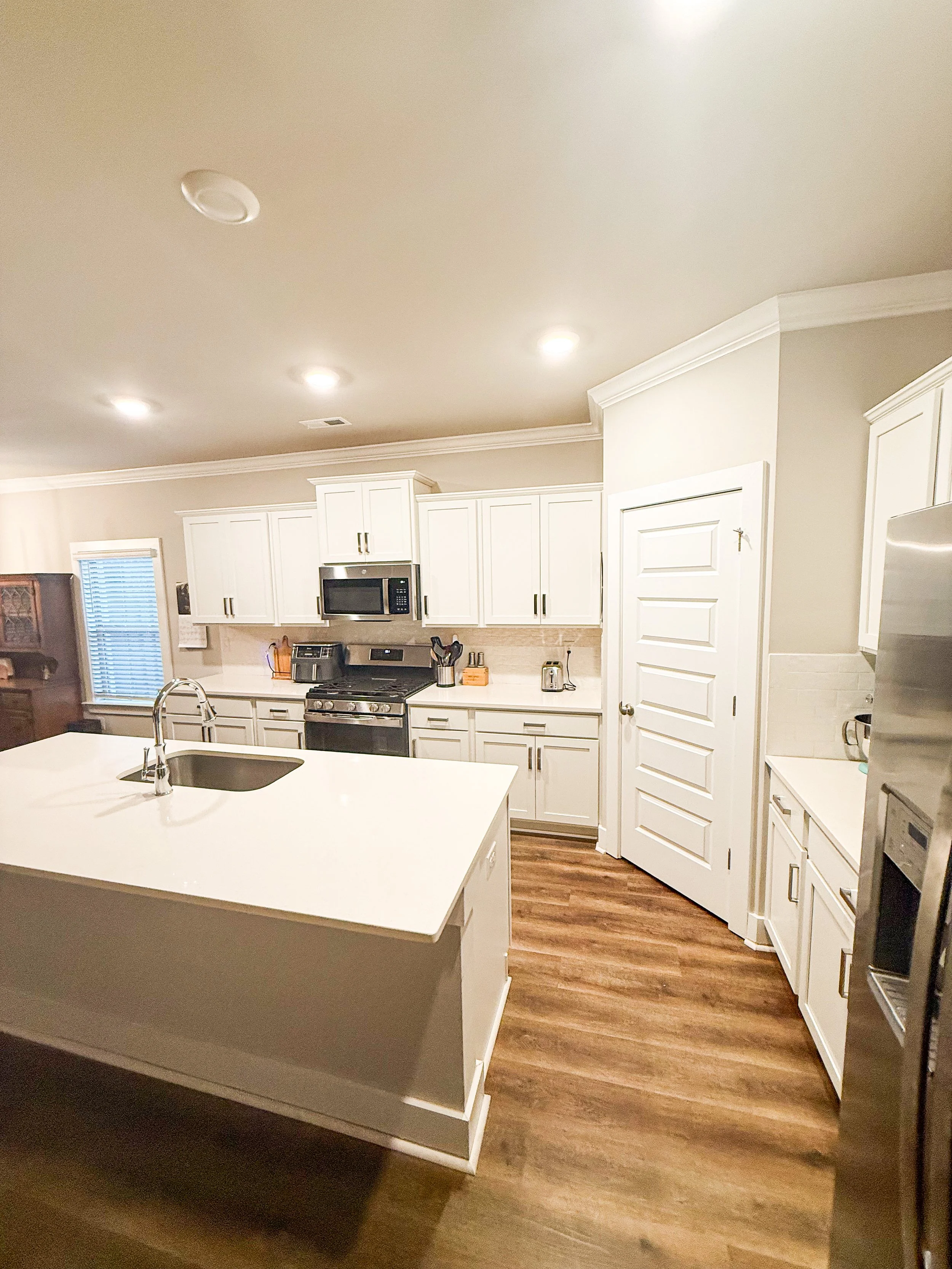 A modern kitchen with white cabinets, stainless steel appliances including a microwave and oven, a kitchen island with a sink, and wooden flooring.