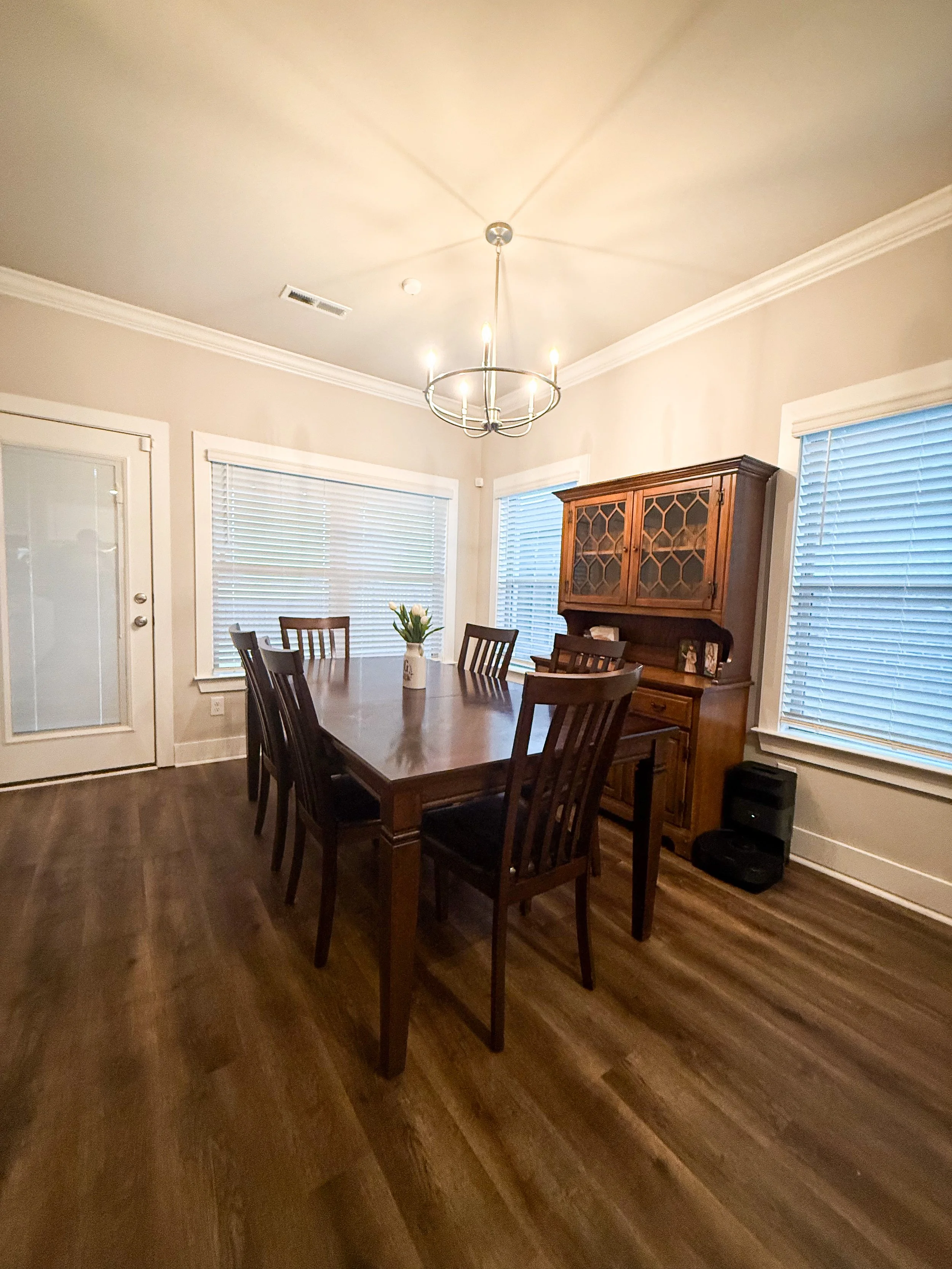 A dining room with a wooden table and six matching chairs, a wooden china cabinet, three large windows with white blinds, a white door with a window, a chandelier with candle-shaped lights, and hardwood floors.