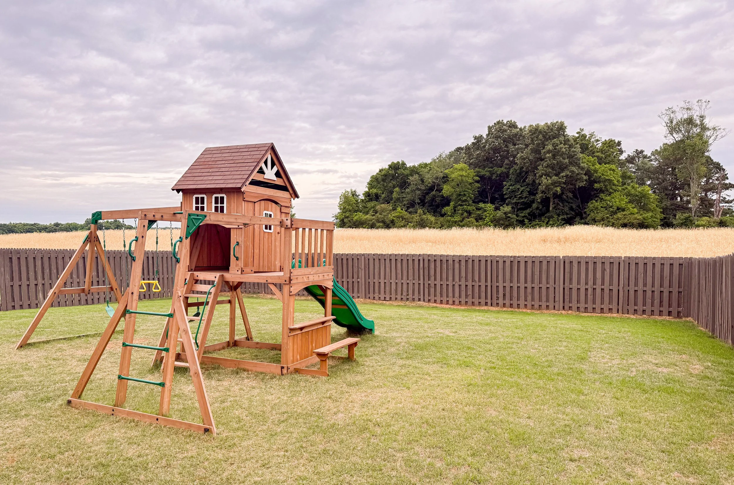 A backyard with a wooden playset that has a small house, swings, and a slide, enclosed by a wooden fence with trees and a field in the background under cloudy sky.