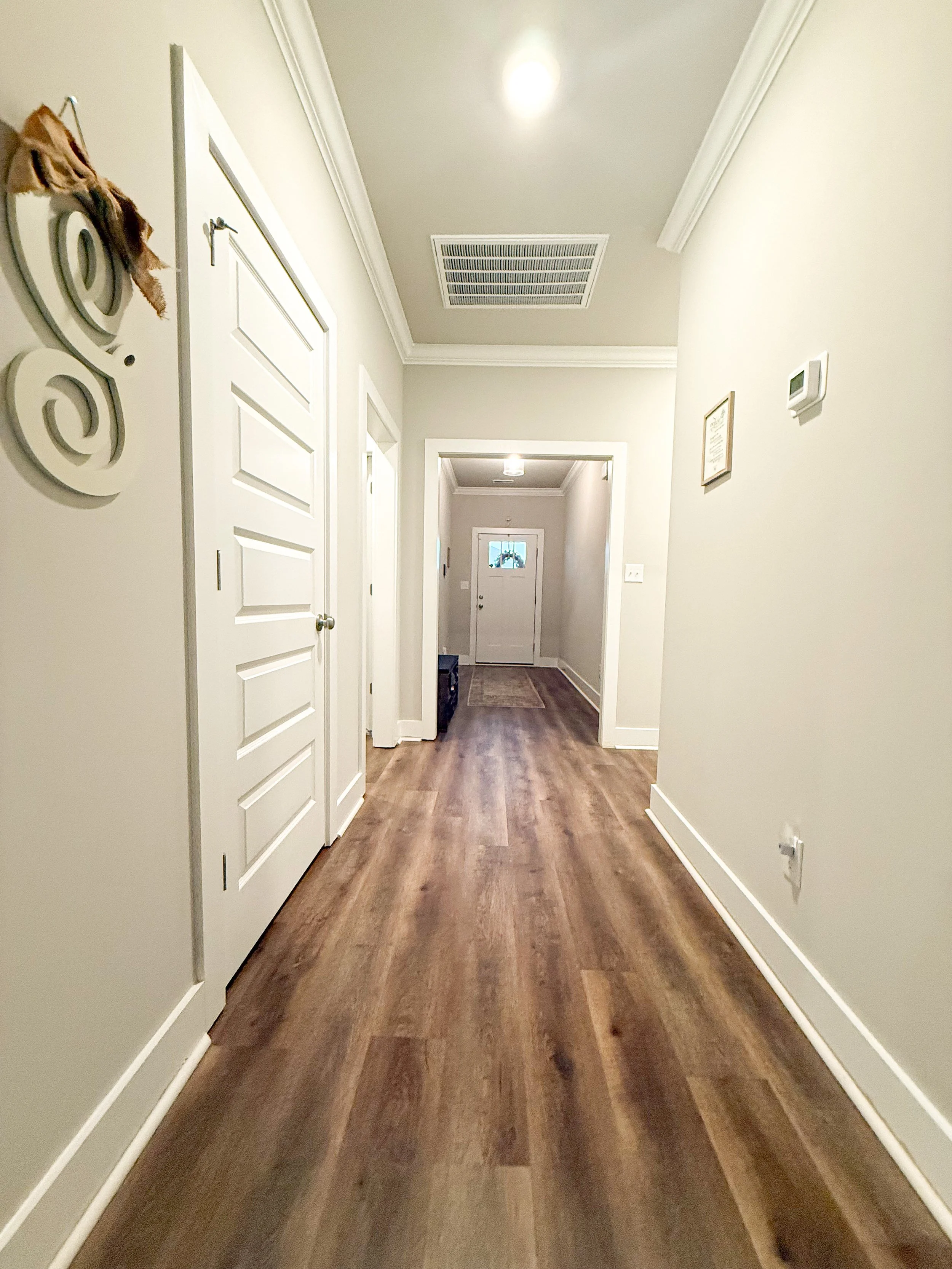 Interior view of a hallway in a house with wood flooring, white walls, and white doors. There are some wall decorations and a framed certificate or document on the right wall. The hallway leads to a closed door at the end.