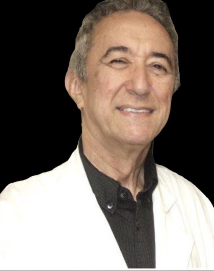 Portrait of a male healthcare professional wearing a white lab coat and a dark shirt, smiling against a black background.