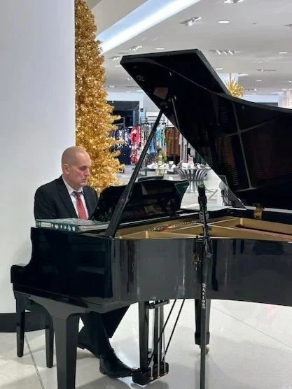 Ron Leonardi in a suit playing a black grand piano inside Nordstrom with holiday decorations.