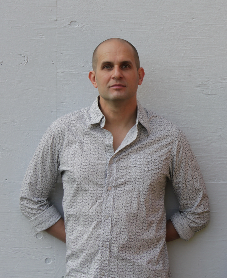 Ron Leonardi with a shaved head and light skin wearing a light patterned button-up shirt standing against a white wooden wall.