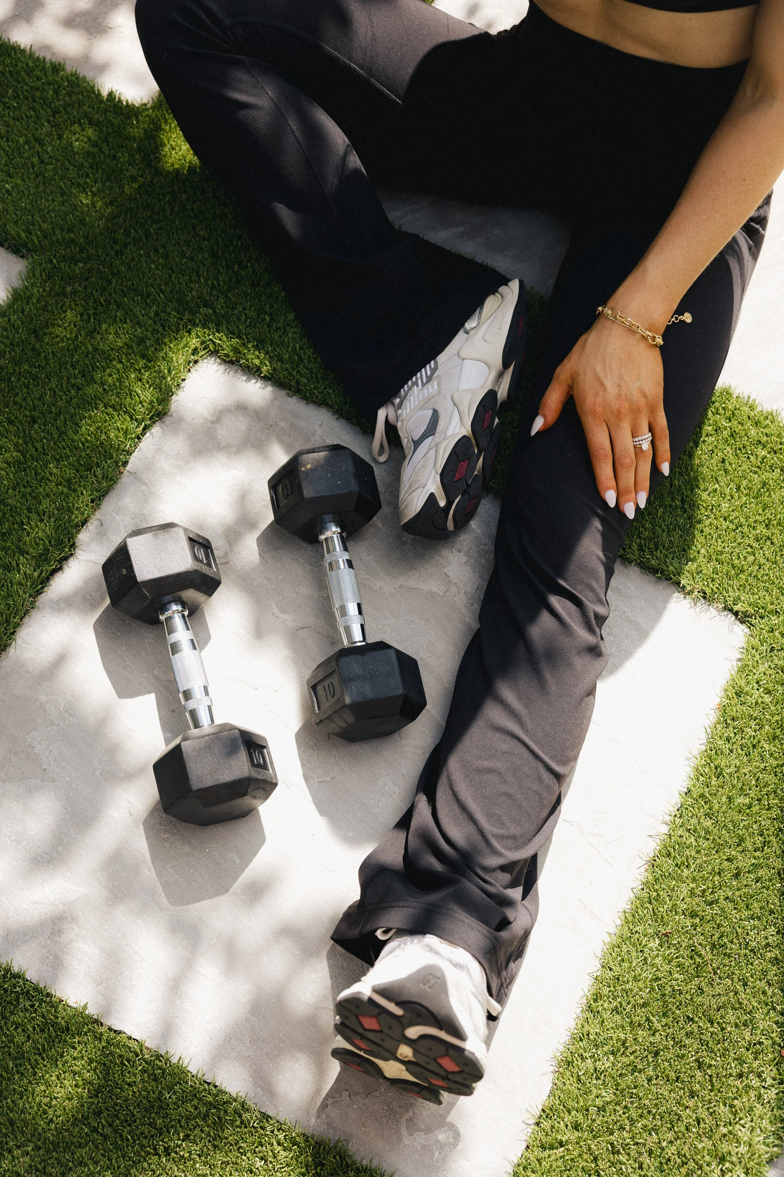 A person sitting on the ground with one leg stretched out and the other bent, wearing athletic shoes, black pants, and a gold bracelet, surrounded by small dumbbells and artificial grass patches on a concrete surface.