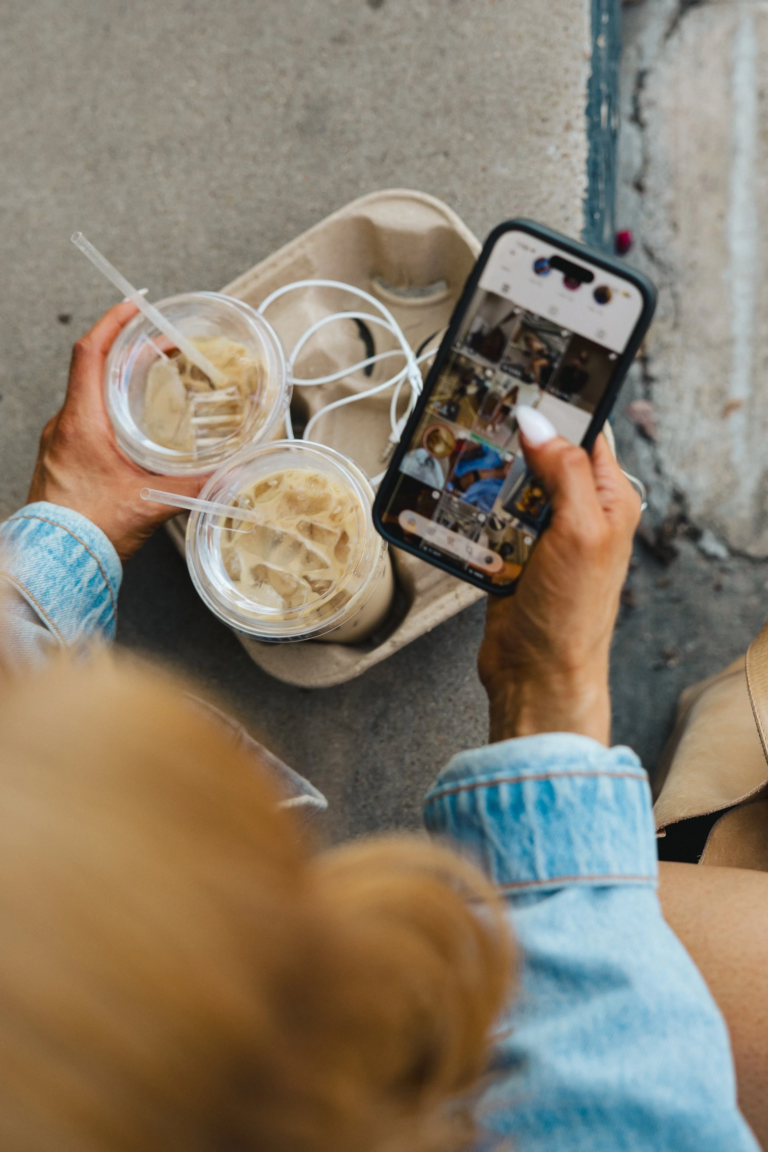 Person holding a smartphone taking a selfie with two iced coffee drinks, while sitting on the ground near a concrete wall.