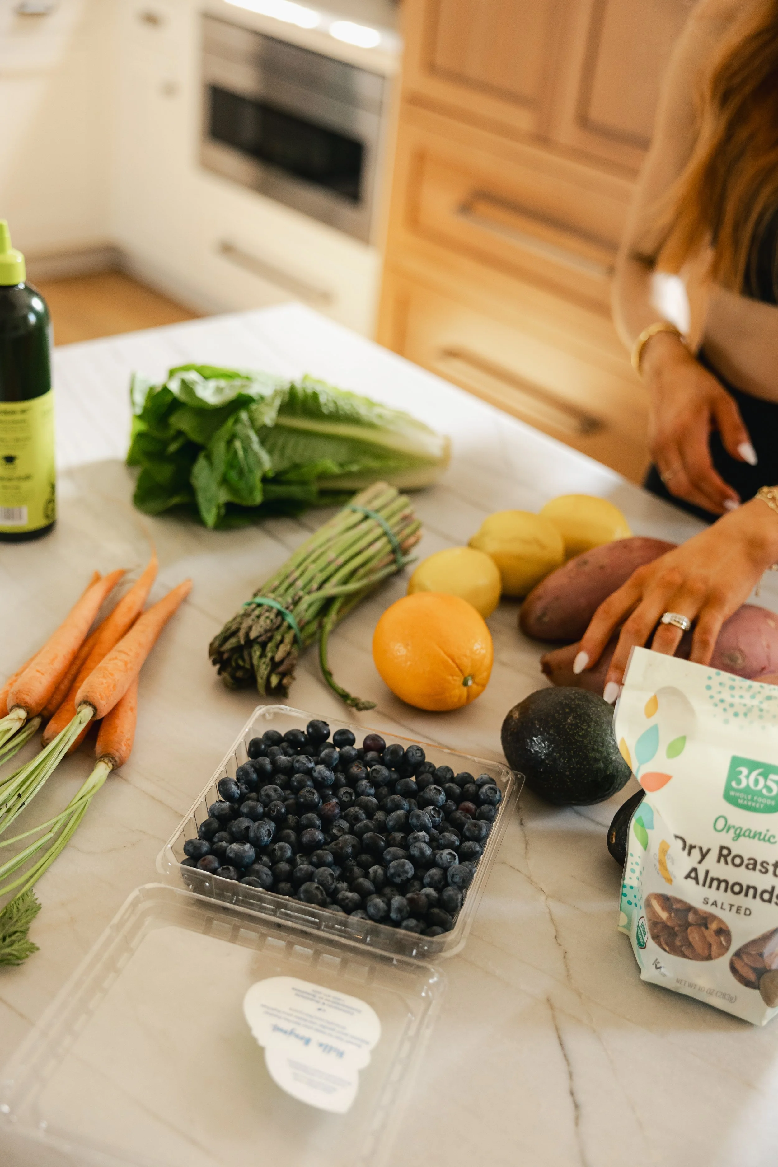 A kitchen countertop with fresh produce including carrots, asparagus, romaine lettuce, oranges, sweet potatoes, an avocado, and a bowl of blueberries. There is also a bottle of green smoothie, a bag of organic dry roasted almonds, and a partially visible woman reaching for food on the right.