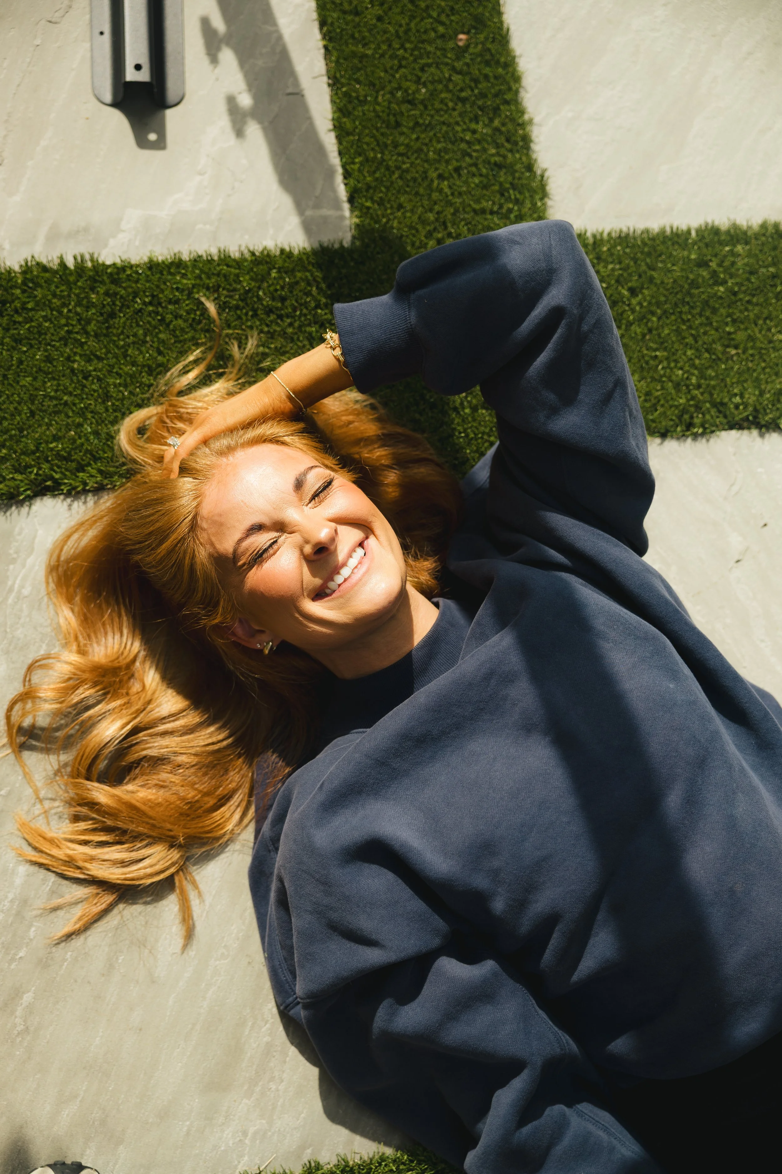 A young woman with wavy blonde hair lying on the ground, smiling with eyes closed. She is wearing a dark sweatshirt, gold jewelry, and has her hand resting on her head. The background includes a patch of green grass and a concrete sidewalk.