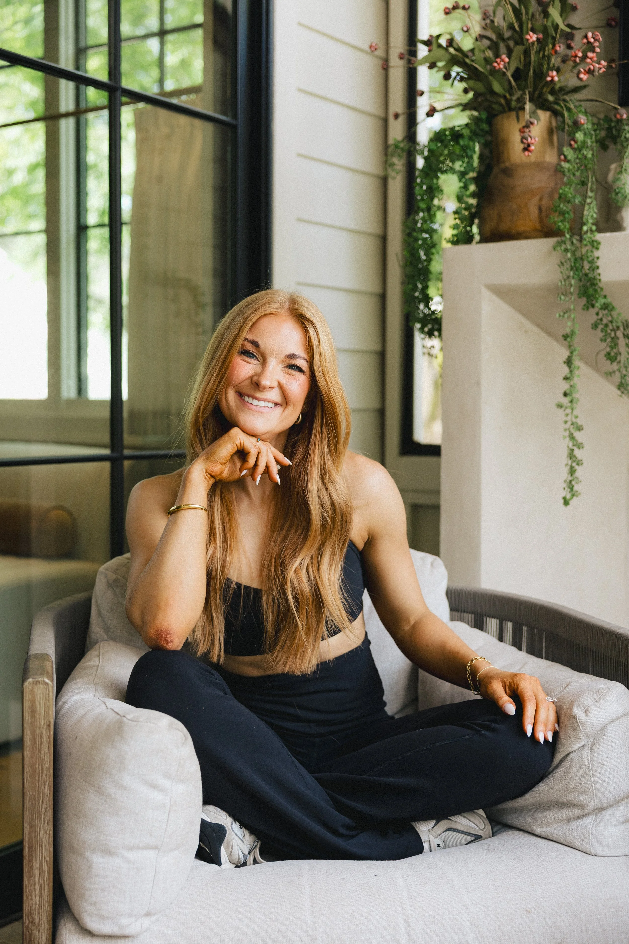 A smiling woman with long red hair sitting on a beige sofa in a bright room with large windows and greenery outside. She is wearing a black crop top and black pants, with jewelry on her wrist and fingers. There is a large plant in a brown vase on a white ledge behind her.