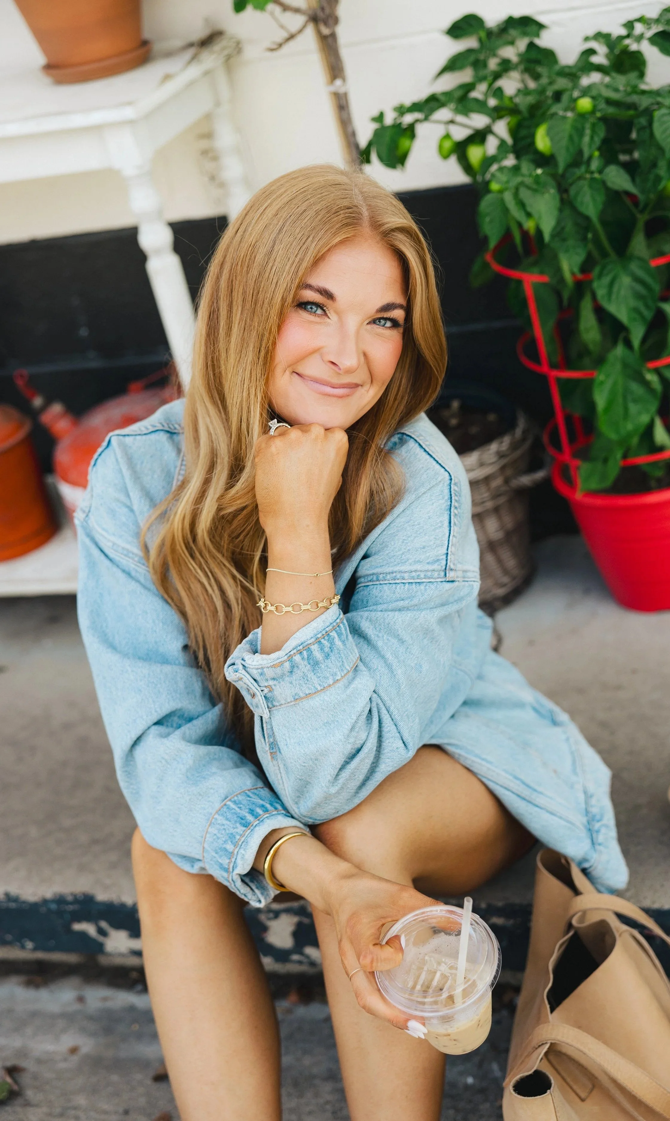 A woman with long blonde hair, blue eyes, and light skin sitting outdoors, smiling at the camera. She is wearing a light denim jacket and jewelry, holding a plastic cup with iced coffee. Behind her, there are potted plants, a white table, and a basket, in a casual, cozy setting.