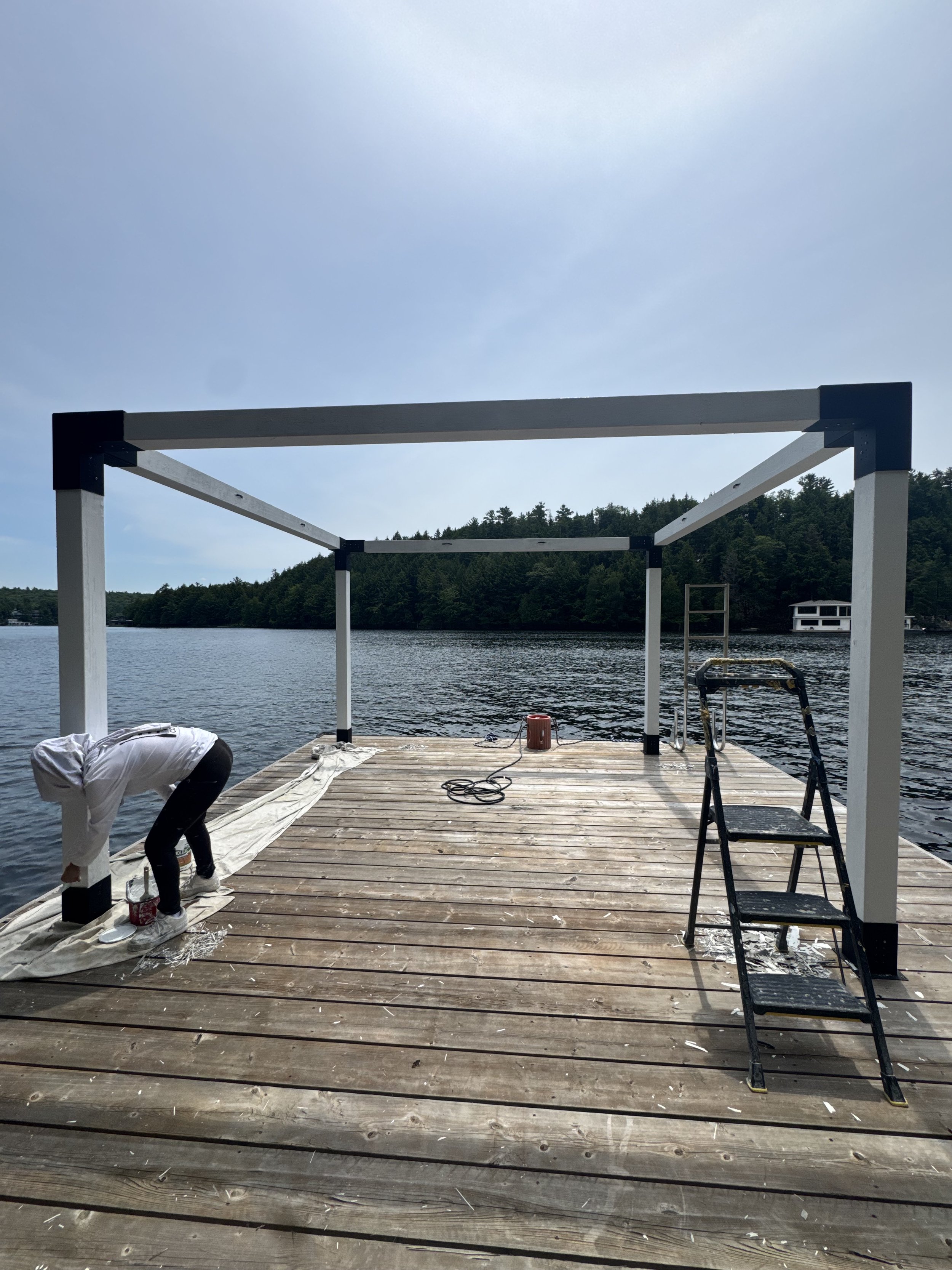 People are working on constructing a wooden dock or platform by a lake, with a partially built white frame structure and a ladder nearby. There are tools, paint cans, and white cloth on the wooden planks. The background features trees, a body of water, and a small white building across the lake.