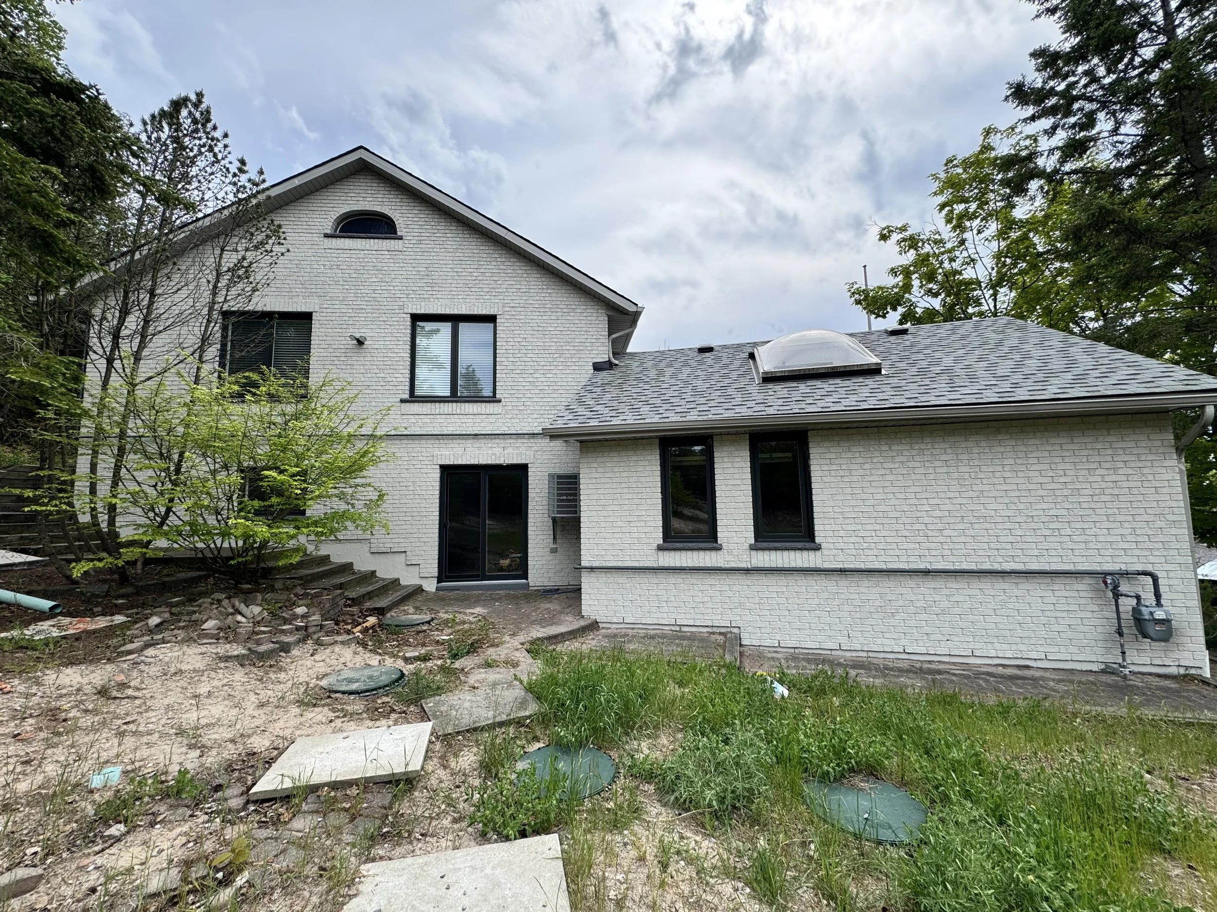 The image shows the back of a white brick house with a gray shingle roof. There are trees around the house, and the sky is cloudy. The yard appears to be under some landscaping or construction, with uneven ground, rocks, and some paving stones.