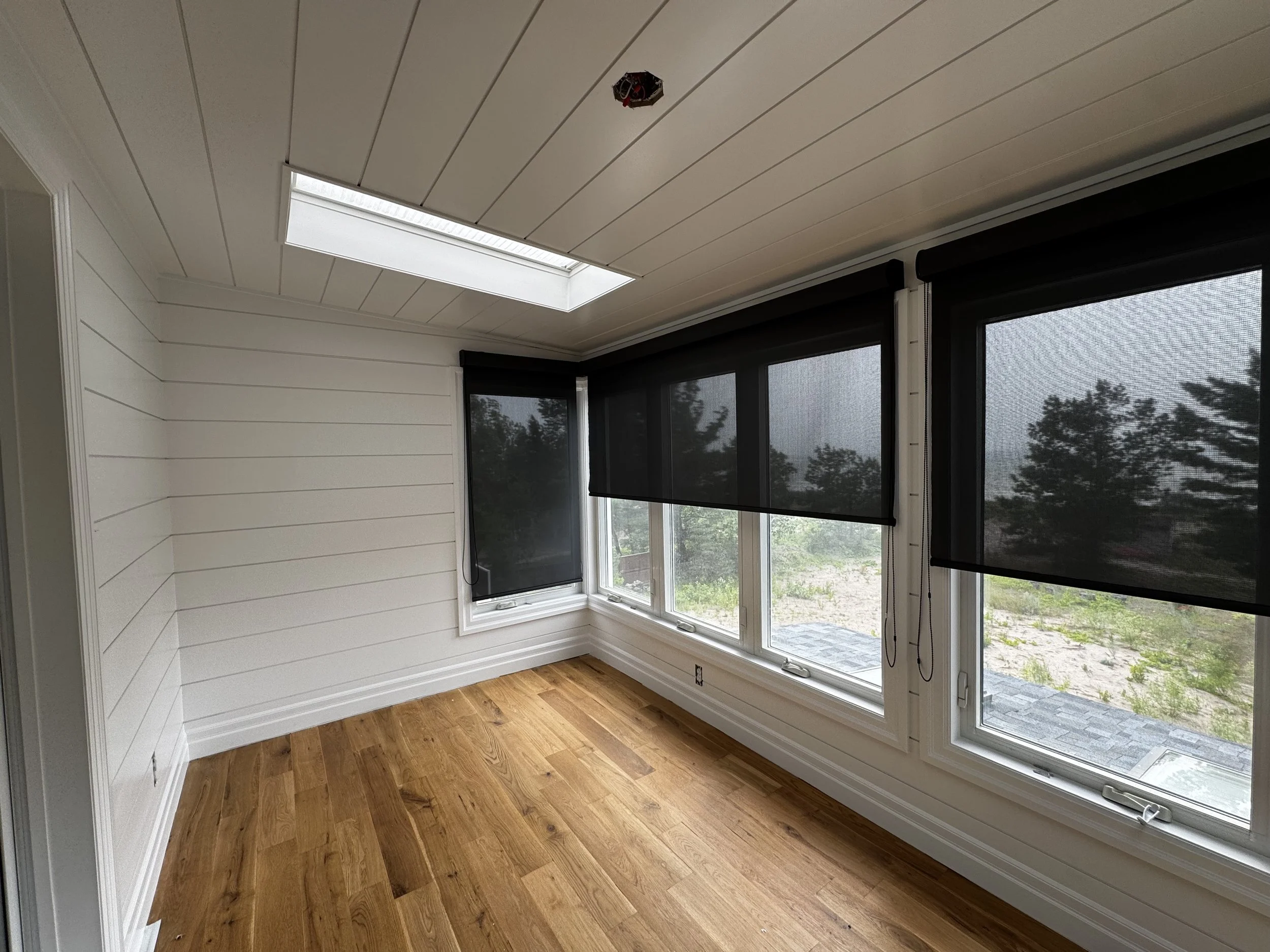 A room with white shiplap walls, a hardwood floor, a window with black roller shades, and a ceiling skylight.