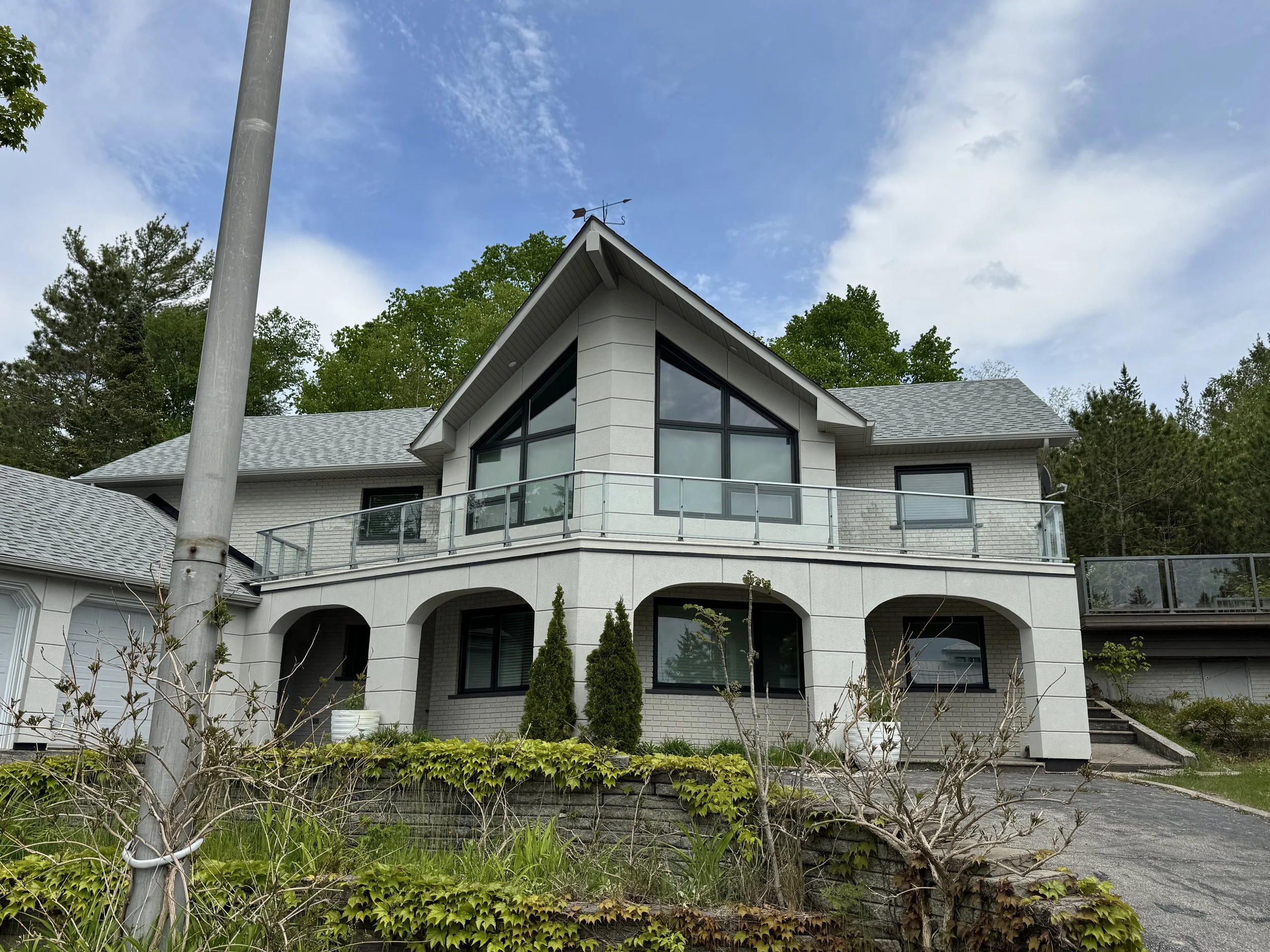 A modern house with large windows is seen behind a yard with green plants and leafless bushes. A tall pole is in the foreground, and trees and a blue sky with scattered clouds are in the background.