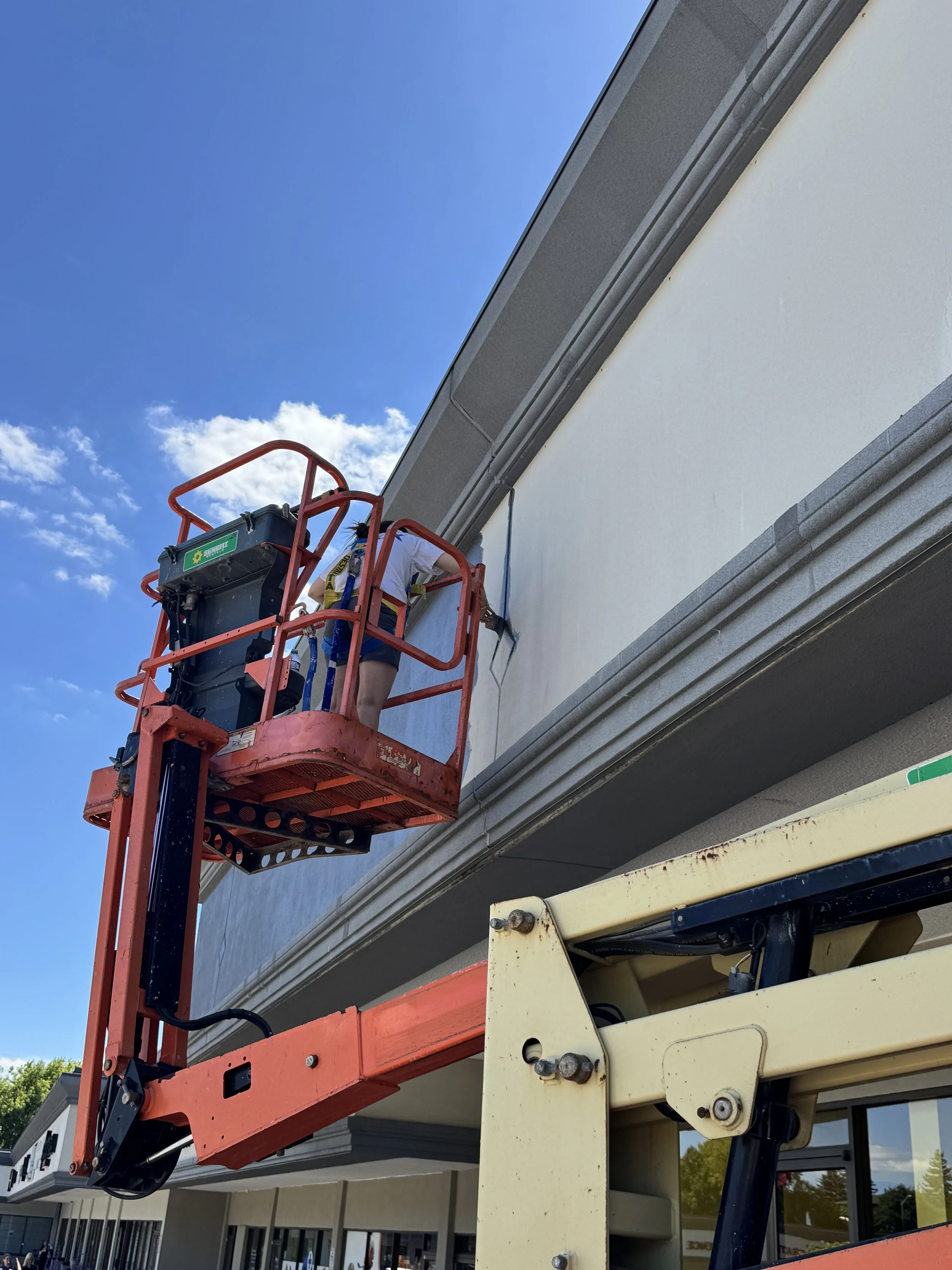 A person operating a cherry picker lift on the exterior of a multi-story building under a blue sky. The individual is working on the building's wall or window area.