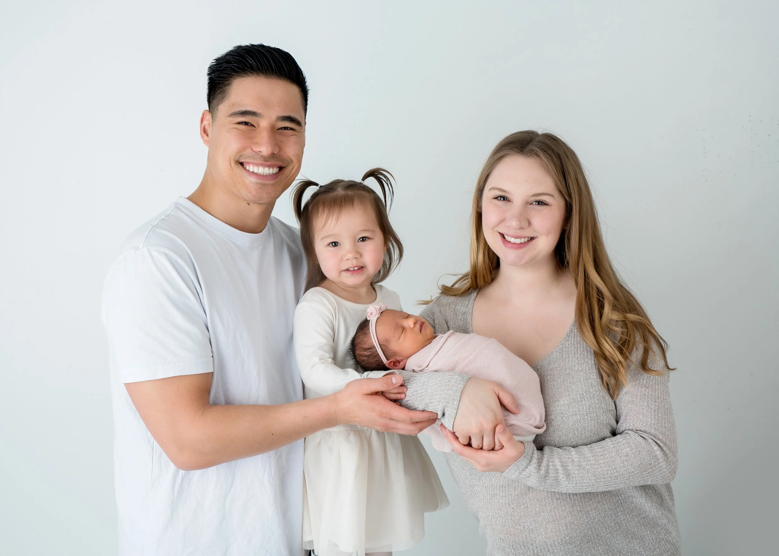 A diverse family of four, including a man, woman, and two young girls, is posing together, smiling, against a plain white background. The woman is holding a newborn baby, and the man is standing next to them.