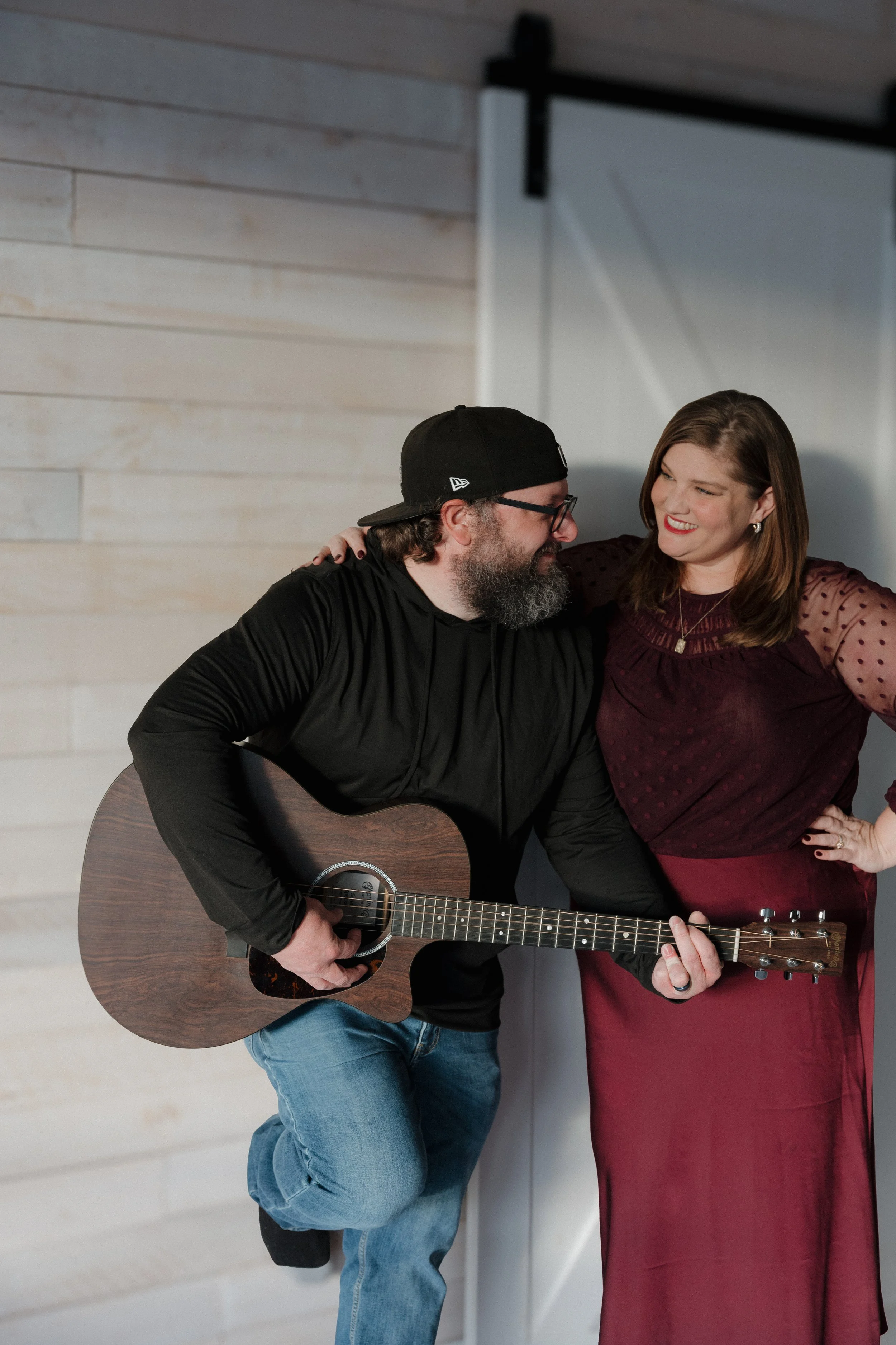 A man playing an acoustic guitar and a woman singing or talking to him, both smiling, indoors with wooden walls.