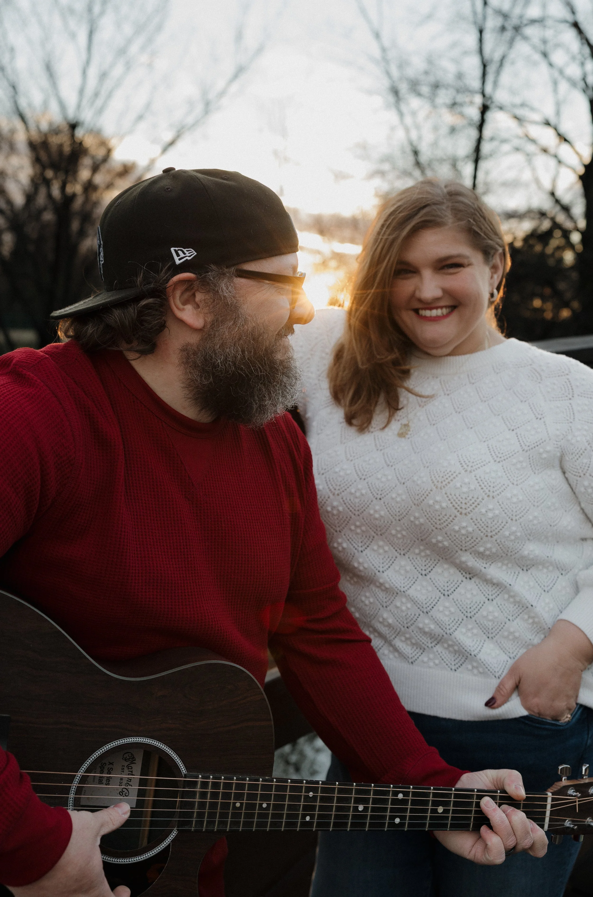A man wearing a black cap, glasses, and a red sweater playing an acoustic guitar, and a woman smiling at him, wearing a white sweater, with trees and a setting sun in the background.