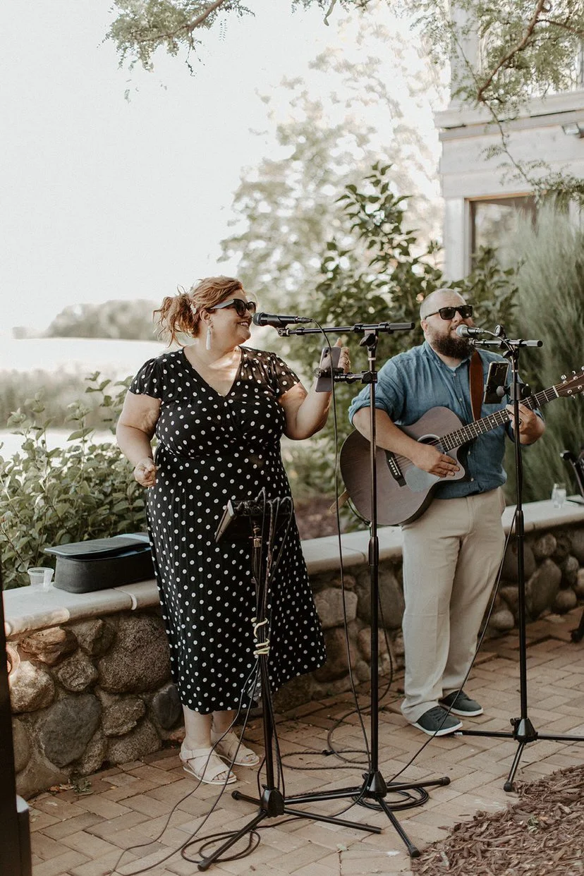 Two musicians, a woman in a black polka dot dress and a man in a blue shirt, are performing outdoors with microphones and a guitar.