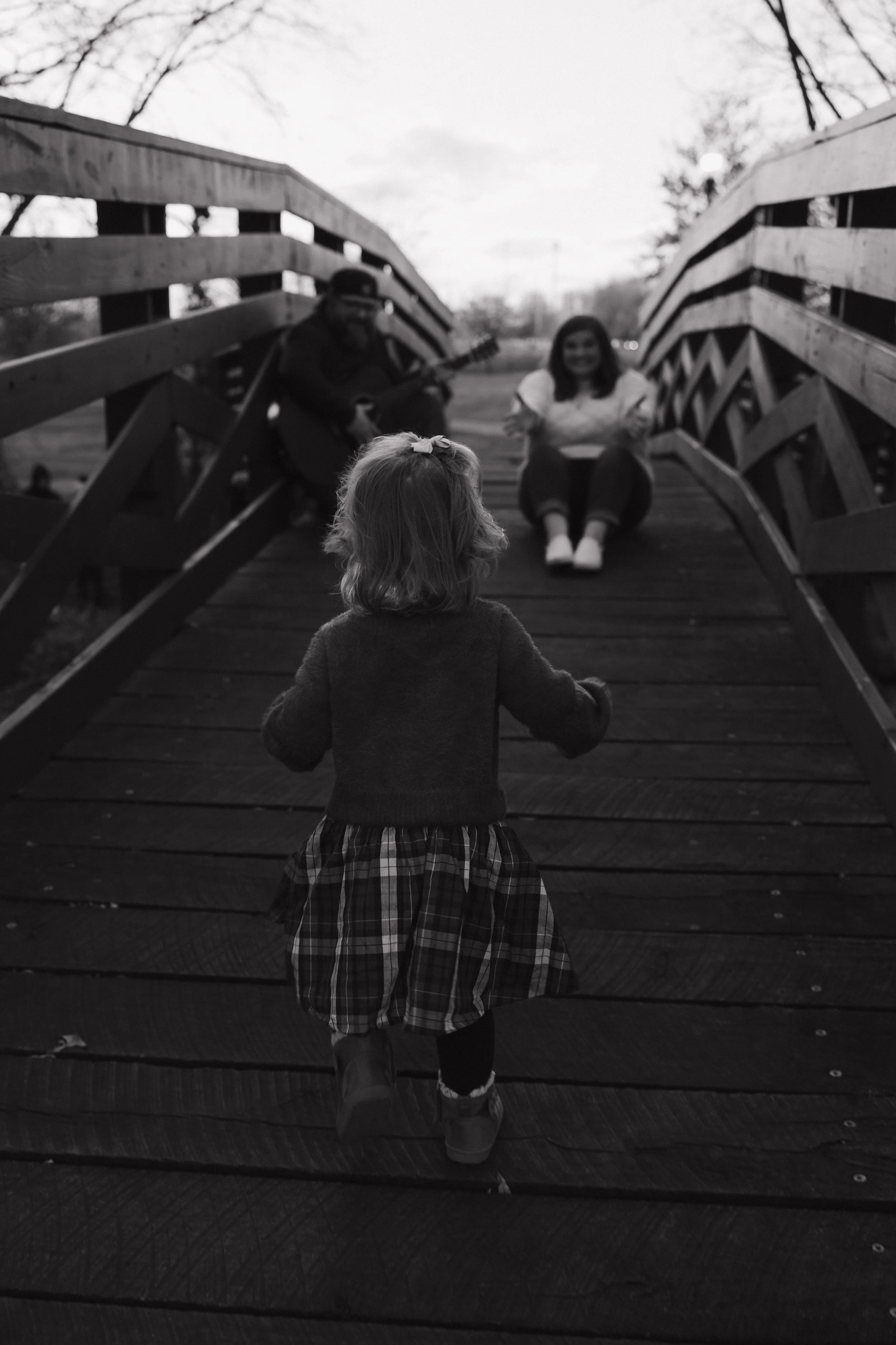 Black and white photo of a young girl walking towards a bridge where two women are sitting, one playing guitar and the other smiling, with trees in the background.