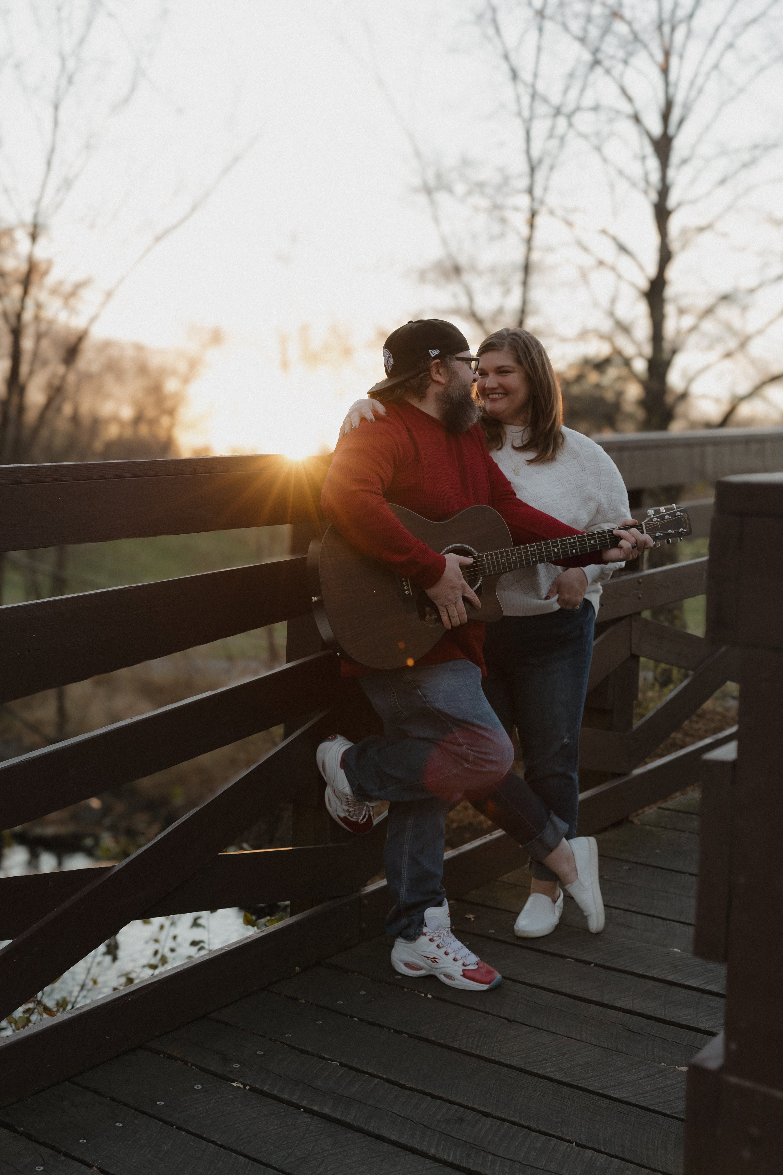 A couple standing on a wooden bridge during sunset, with the man playing guitar and the woman smiling, in a park with trees in the background.