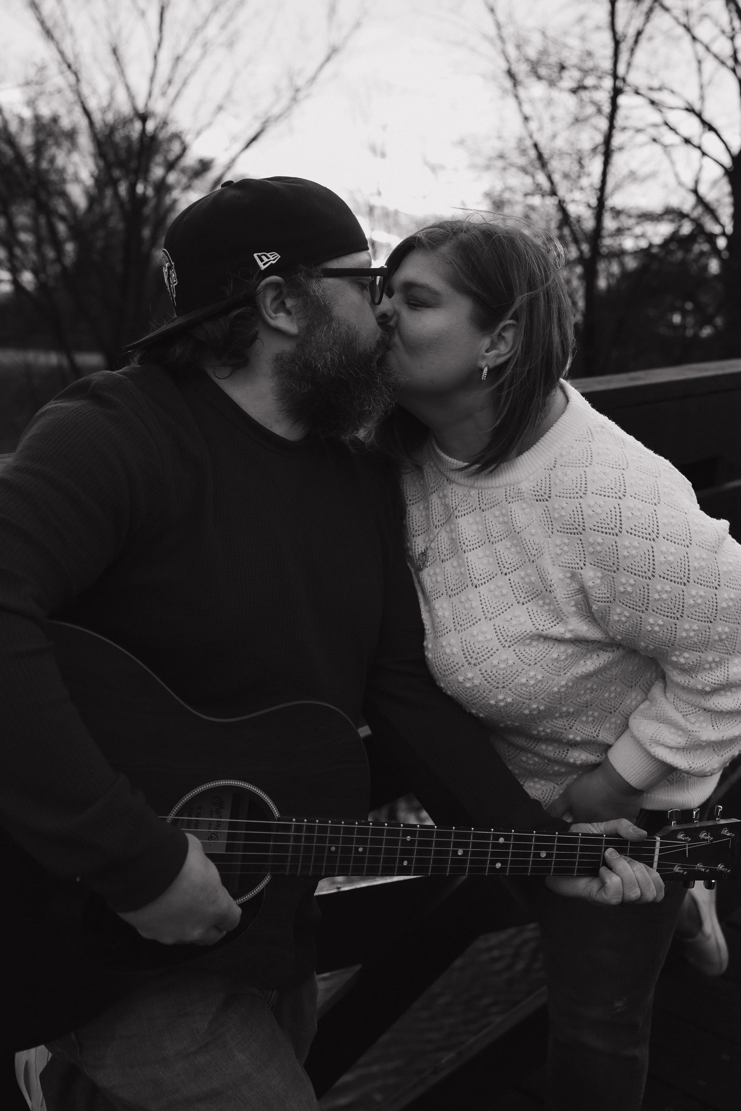 A grayscale photo of a man and woman sharing a kiss outdoors, with trees in the background. The man is wearing a baseball cap and glasses, and the woman has earrings and a patterned sweater. The man is also holding a guitar.