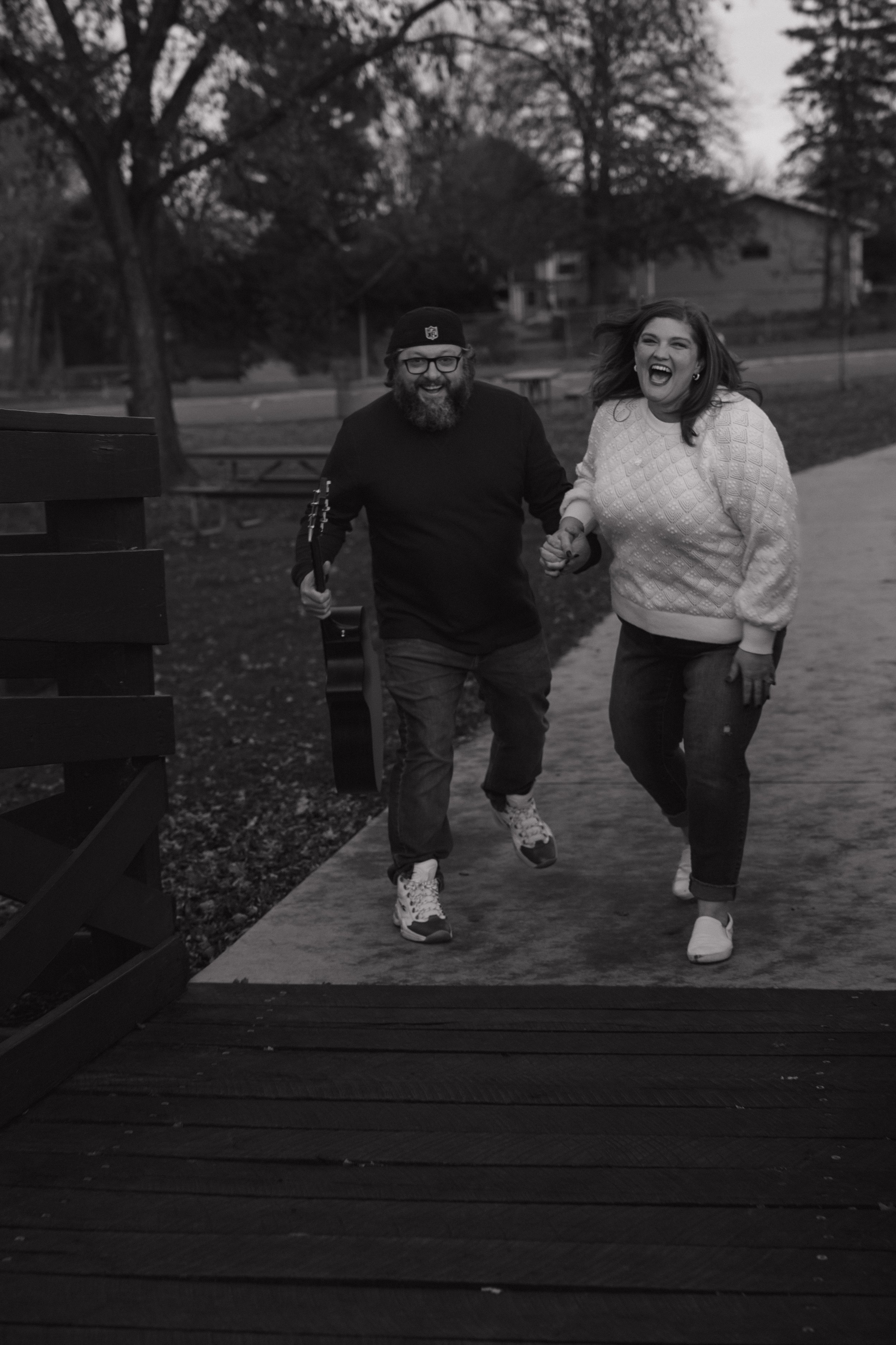A man and woman are walking hand in hand, smiling and laughing outdoors on a sidewalk with trees and houses in the background. The man is wearing glasses, a beanie, and a dark sweater, holding a guitar. The woman is wearing a light-colored knit sweater and white shoes.