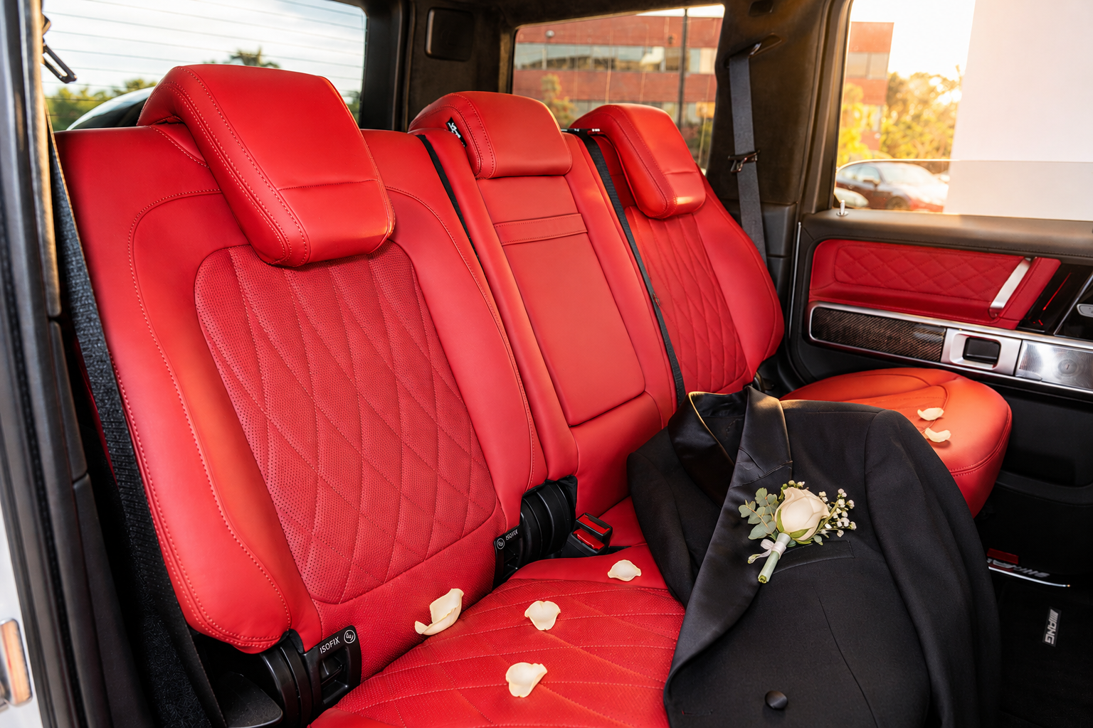 Red leather upholstery inside a Mercedes-Benz G63 with wedding roses and flower petals on the seats, black jacket with boutonniere on the seat, and a view of the outside through the window.